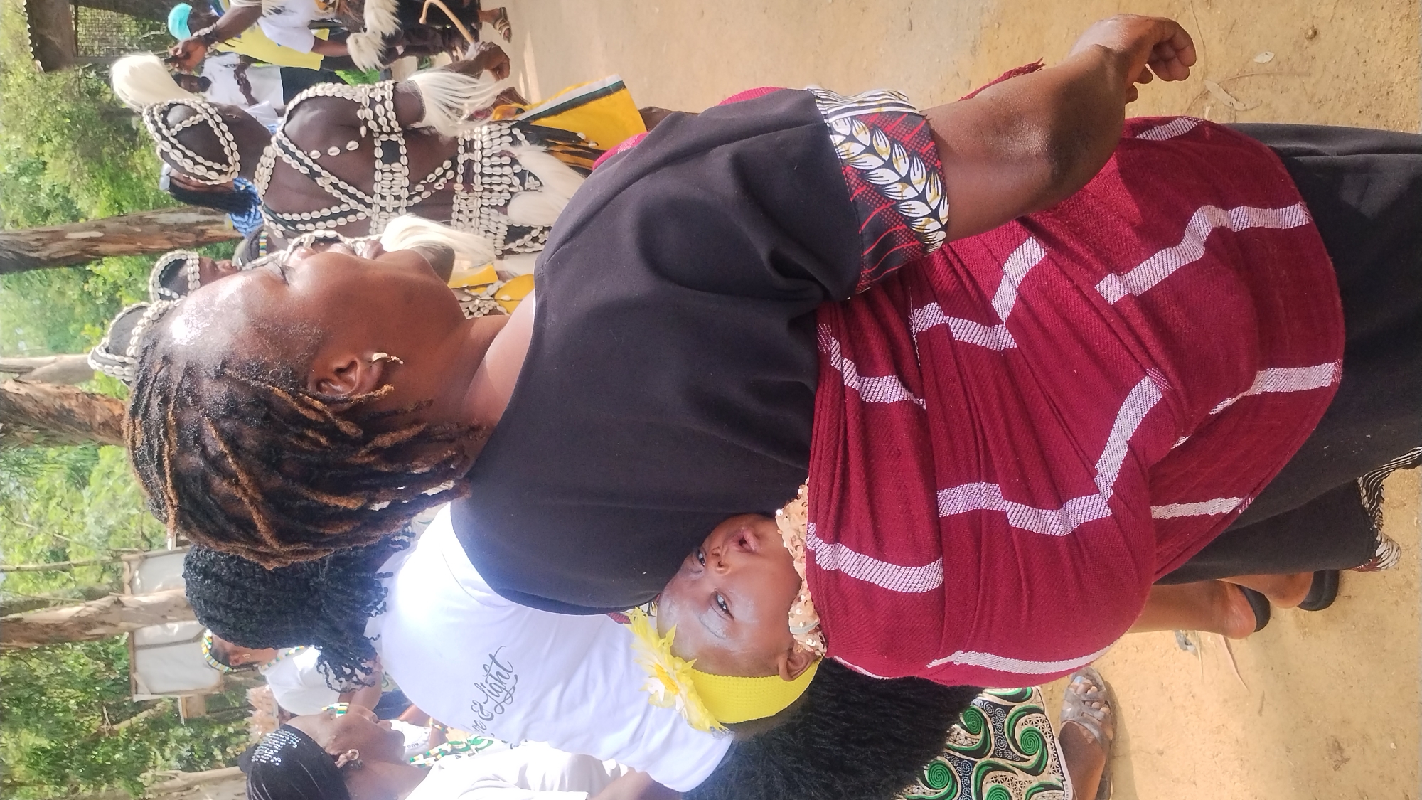 In Jos, Nigeria, a woman carries a baby on her back, wrapped in a maroon and white patterned cloth. The woman, with dreadlocks and wearing a black top with patterned cuffs, faces away from the camera and to the right, smiling. The baby, wearing a yellow headband with a flower, is visible over the woman's shoulder. The background suggests an outdoor gathering with trees, other people in traditional attire, and what appears to be a makeshift structure. The ground is dirt.
