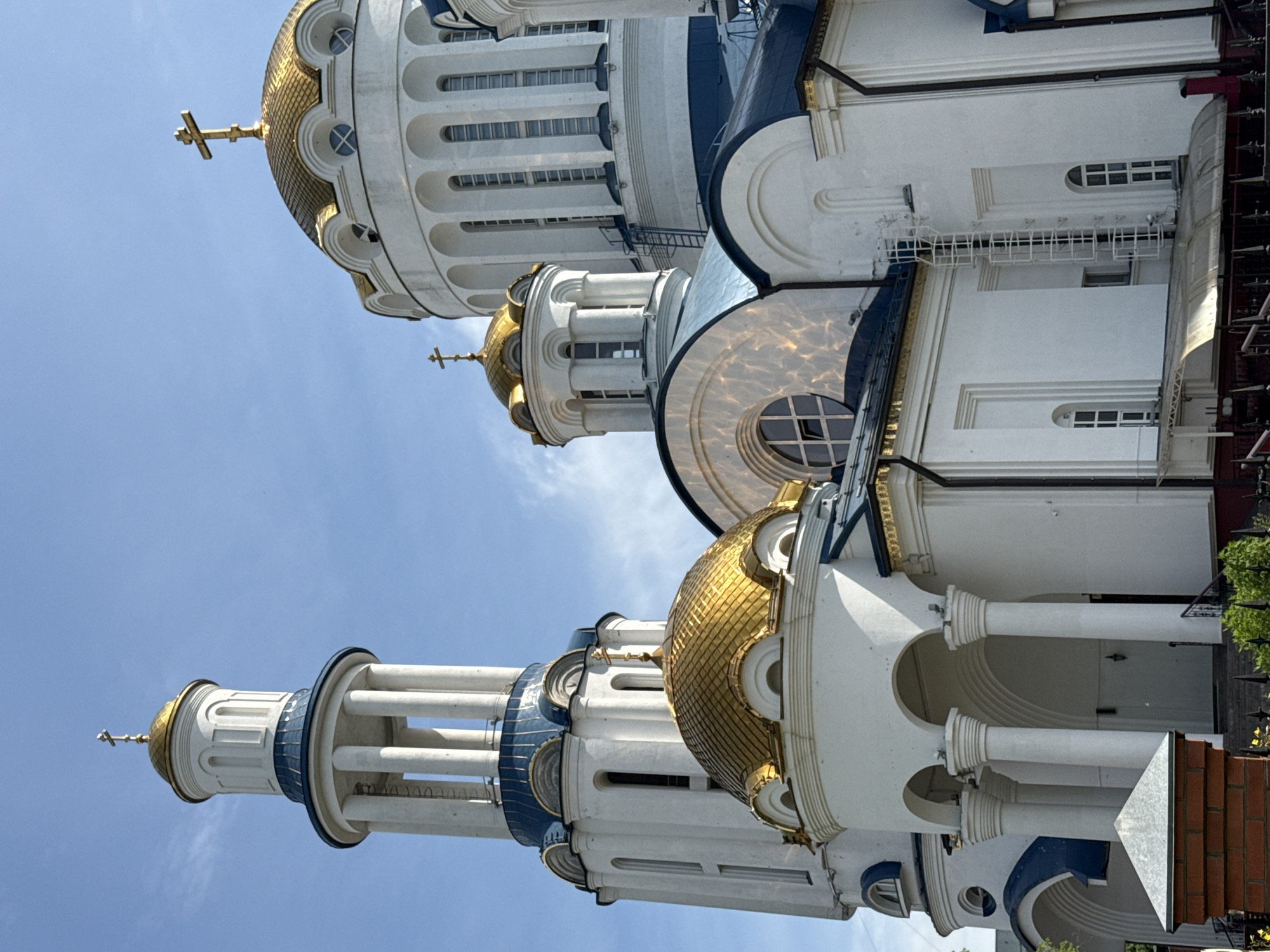 The image shows the Church of the Ascension in Izmaylovo, Moscow, Russia. It is a white, multi-domed Orthodox church with golden domes and blue accents. The church has a tall bell tower and several smaller domes. A cross is visible atop the tallest dome. The sky is blue with some wispy clouds. The lighting suggests it is daytime.
