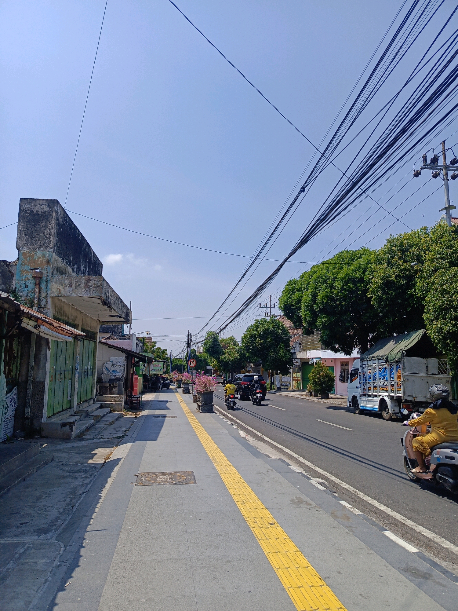 The image depicts a street scene in Magetan, Indonesia, on a clear day with a bright blue sky. A sidewalk runs along the left side of the street, featuring a textured yellow tactile paving strip for the visually impaired. Several shops with green, weathered facades and rusty awnings are visible along the sidewalk. One building has a prominently crumbling concrete structure at the top.
The street itself is paved and carries moderate traffic. A white truck with a green tarp over its cargo is seen driving in the right lane. Several motorcycles are also present: one ridden by a person in a yellow outfit is in the foreground on the right, and others are further down the road. A black car is also visible in the traffic. Lush green trees line the right side of the street, with some houses interspersed among them. Overhead, a dense network of dark electrical wires crisscrosses the sky, supported by poles. The scene appears to be a typical urban commercial or residential area.