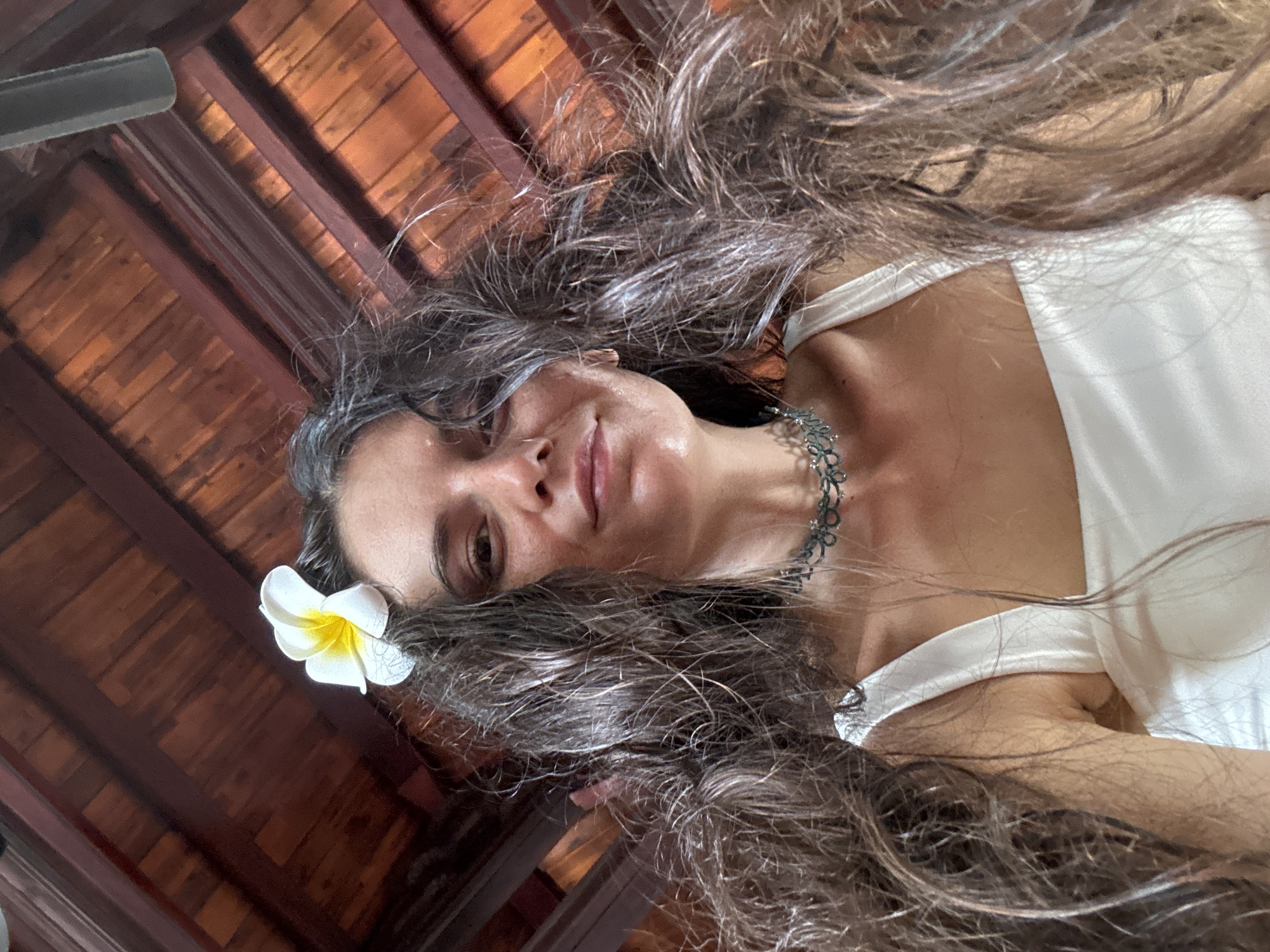 A close-up, low-angle selfie shows a woman with long, dark, wavy hair, appearing to lie down and look upwards at the camera. A white plumeria flower with a yellow center is nestled in her hair above her left ear. She is wearing a white top, possibly a tank top or camisole, and an intricate dark choker necklace. Her expression is calm and serene, with a faint, gentle smile.
The background features a wooden ceiling composed of dark beams and lighter wooden planks, indicating a rustic or tropical architectural style. A dark, blurred object, likely a ceiling fan blade, is visible in the upper left section of the image. The combination of the wooden interior and the plumeria flower suggests a tropical resort or a warm climate setting, consistent with the provided location context of Mũi Né, Vietnam. There is no visible text in the image.