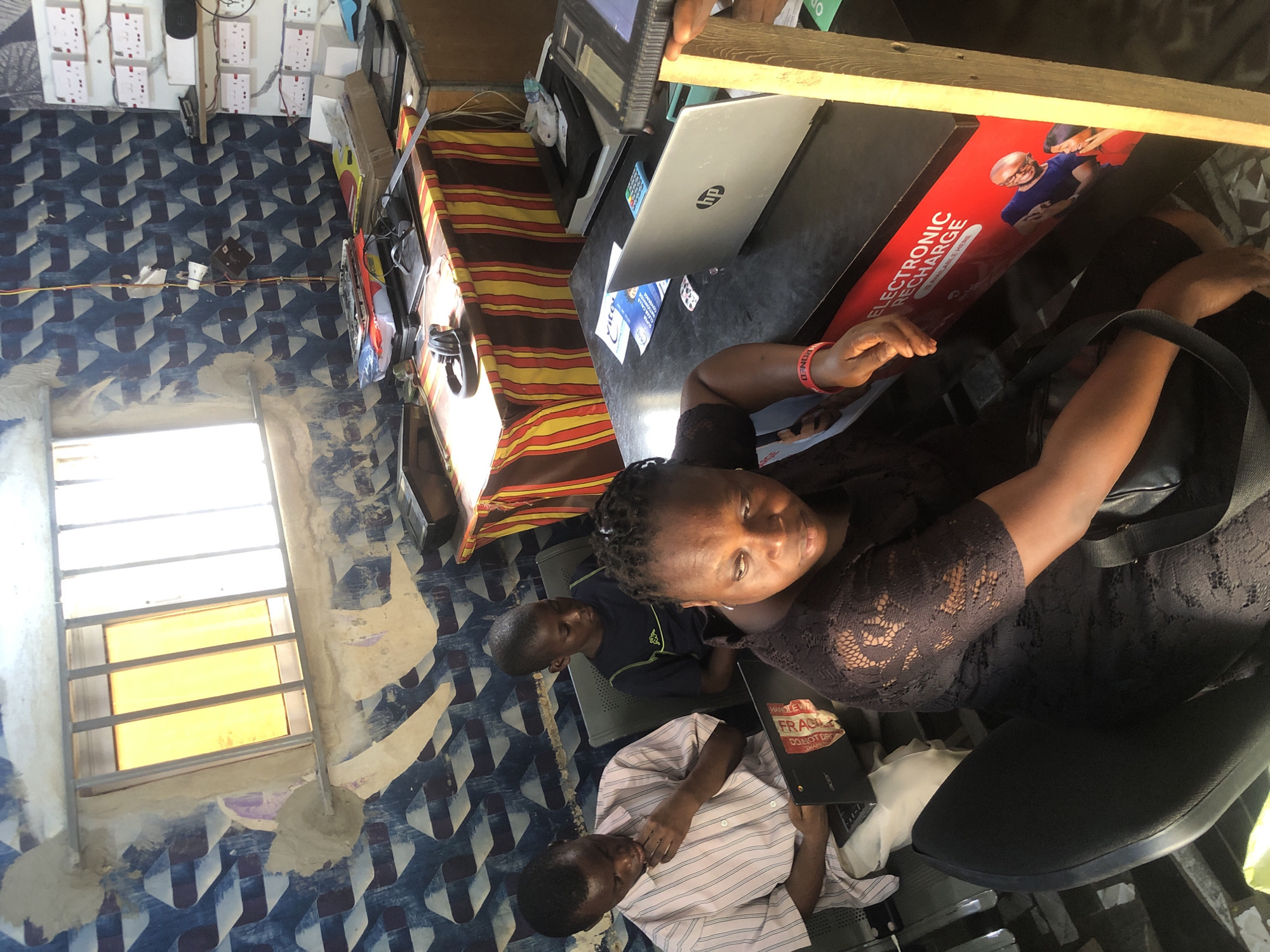 In the city of Ado, Nigeria, this image captures an indoor scene, possibly a small business or office. A woman in a black lace dress is seated in the foreground, her expression downcast. Beside her, a man in a striped shirt is looking down at a laptop. A young boy in a blue t-shirt sits behind them, also looking down. The room is decorated with a blue and white patterned wallpaper and features a desk area with laptops, a printer, and various electronic devices. A red sign on the desk reads "ELECTRONIC RECHARGE IS AVAILABLE HERE." A window with metal bars is visible in the background, with light streaming through. The overall impression is one of a busy, functional space, perhaps a cyber cafe or an electronics repair shop.
