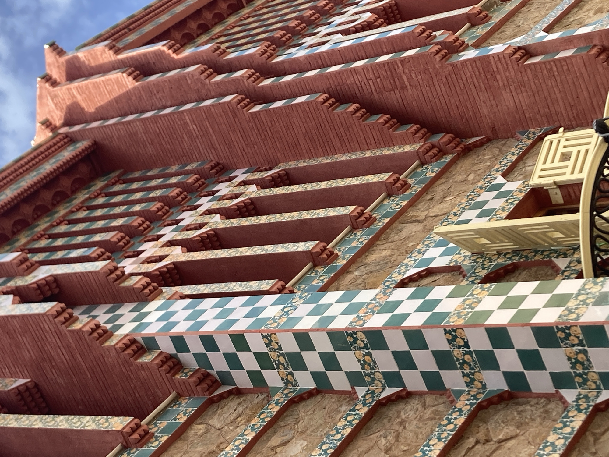 The image shows a detailed view of the facade of Casa Vicens, a modernist building located in the Gràcia district of Barcelona, Spain. The facade is characterized by its extensive use of red brick and decorative ceramic tiles. A prominent feature is a column covered in a checkerboard pattern of green and white tiles. Above this, a band of tiles with a floral pattern in yellow and green runs horizontally. The brickwork of the building exhibits a textured, almost ribbed, appearance. The windows are framed by decorative elements, with vertical bands of alternating white and light blue tiles interspersed with the red brick. The architectural style is distinctively Catalan Modernism. The sky is visible in the upper right corner, with some light clouds. A portion of a wrought-iron balcony and a decorative wooden shutter are visible in the lower part of the image.