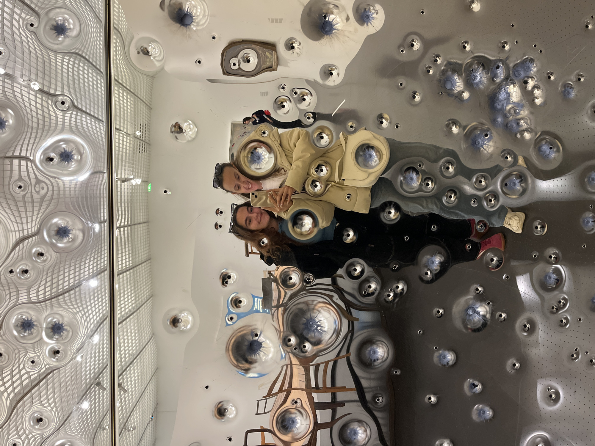Two women are taking a selfie in a reflective installation in Metz, France. The installation is characterized by its mirrored surface adorned with numerous convex protrusions, creating distorted reflections of the surrounding environment and the women themselves. The background includes a window with a grid-like pattern. The women are positioned closely together, looking at the camera, with one holding a phone. The reflections show additional elements of the installation and the environment.