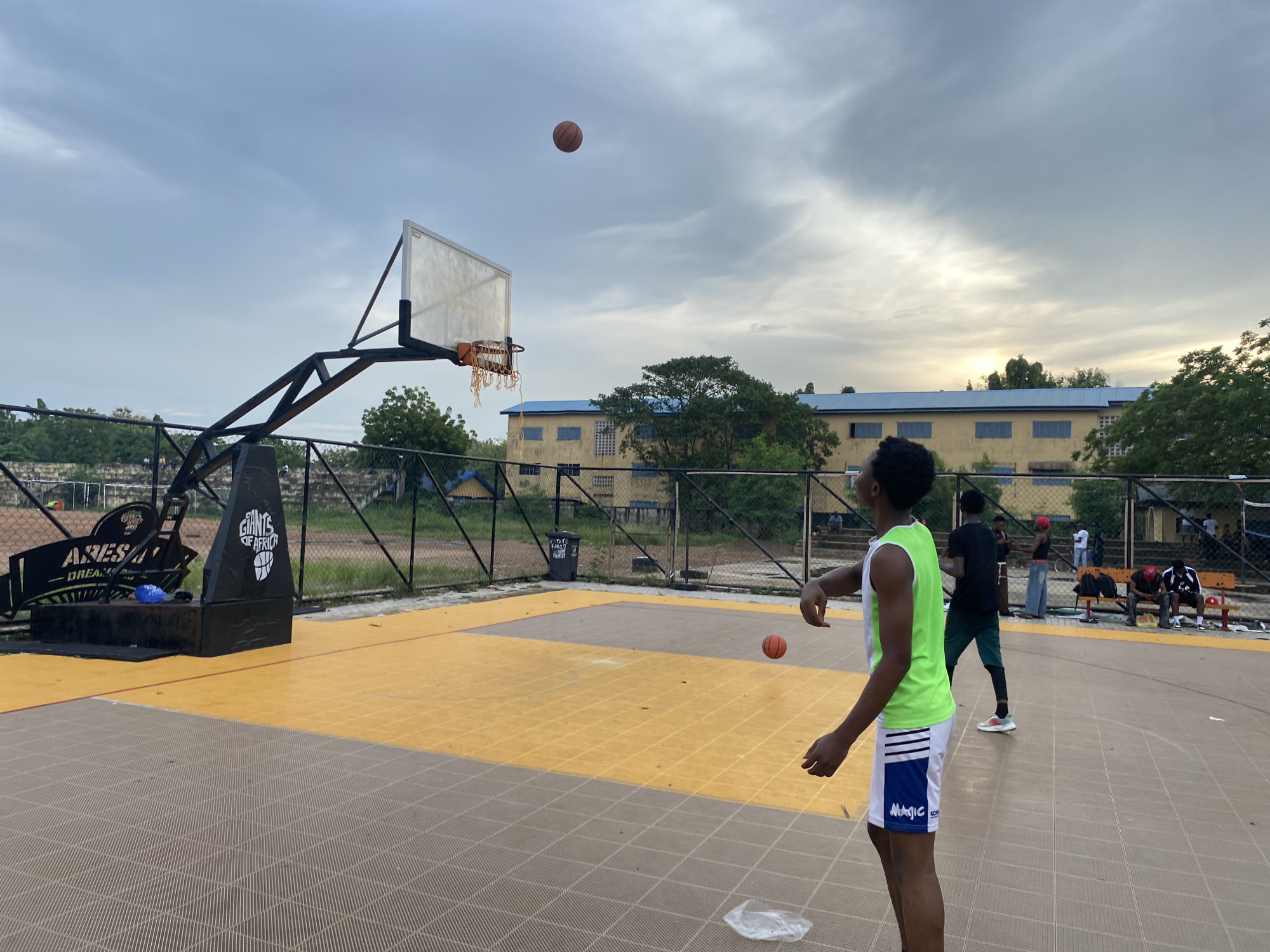 A medium shot captures a youth throwing a basketball at a basketball court in Lagos, Nigeria. The court has a gray-and-yellow pattern. The youth faces left, wearing a white tank top with a green border and white-and-blue shorts. In the background, another person stands still. Behind the basketball court, a metal fence separates the court from a building with multiple windows and a blue roof. A basketball hovers in the air above the basket and to the side, a plastic bag sits on the ground. People are sitting on benches further back on the right. The sky is overcast, and the scene appears to be taken in the late afternoon.
