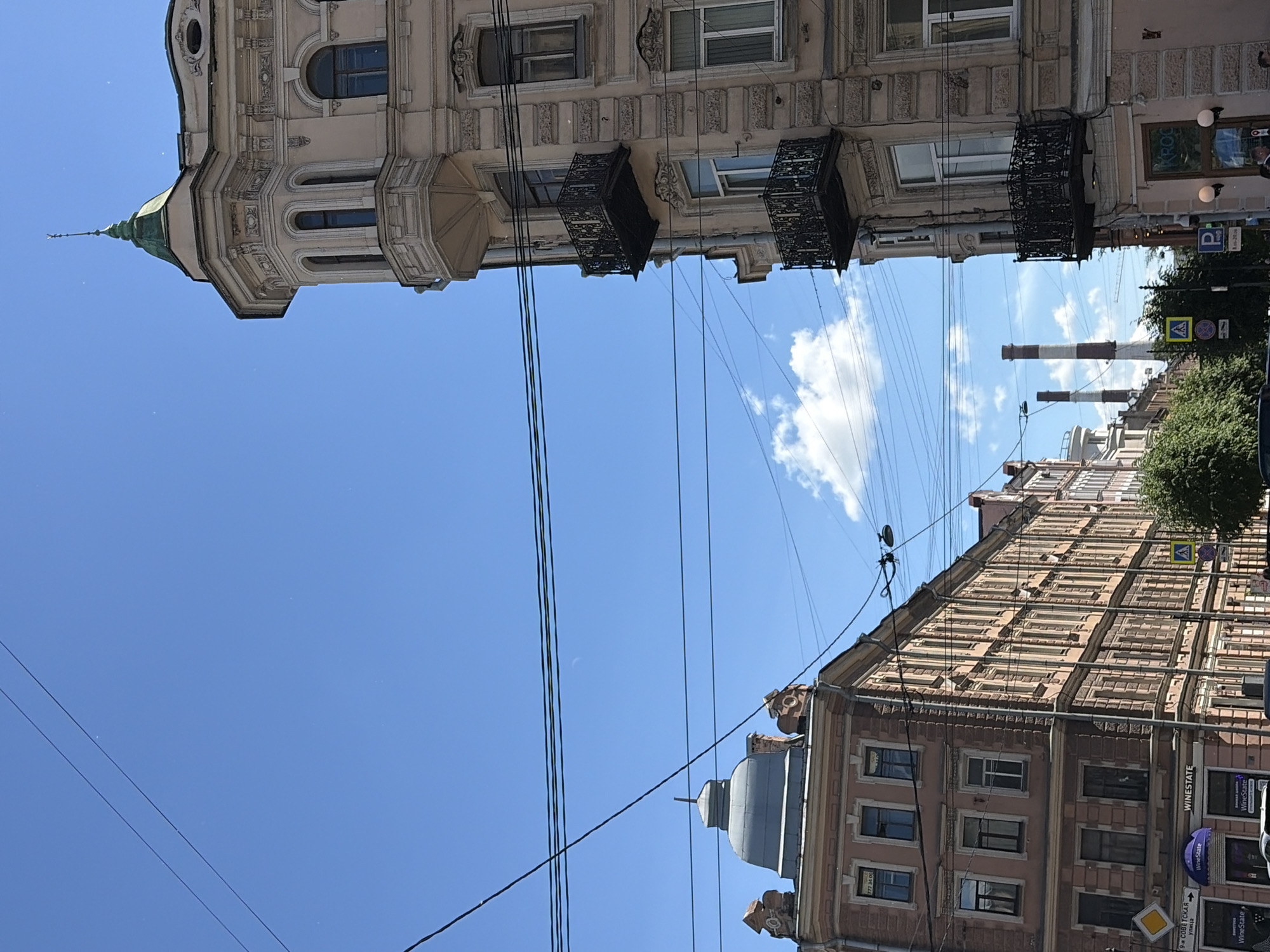 The image captures a street scene in Saint Petersburg, Russia, under a bright, clear blue sky with scattered white clouds. Two tall buildings dominate the foreground. The building on the right, a pale peach color, features an ornate corner with a conical green-capped turret, multiple arched windows, and decorative balconies with intricate black metal railings. It exhibits classic European architectural style.
To the left, a contrasting building with a grey domed roof and a similar peach-colored facade stands. This building also has multiple windows and decorative architectural elements. Further down the street, a long row of buildings with many windows extends into the background, displaying a consistent architectural style.
Overhead, a complex network of electrical wires crisscrosses the sky, typical of urban environments. In the distance, two tall industrial chimneys with red and white bands are visible, suggesting a nearby industrial area or power plant. Several street signs, including traffic signs and directional arrows, are present on the sidewalk. A few trees with green foliage line the street, adding a touch of nature to the urban landscape. There are no visible people actively engaged in any specific actions in the foreground. The overall impression is a sunny day in a historic city, with a blend of old-world architecture and modern infrastructure.