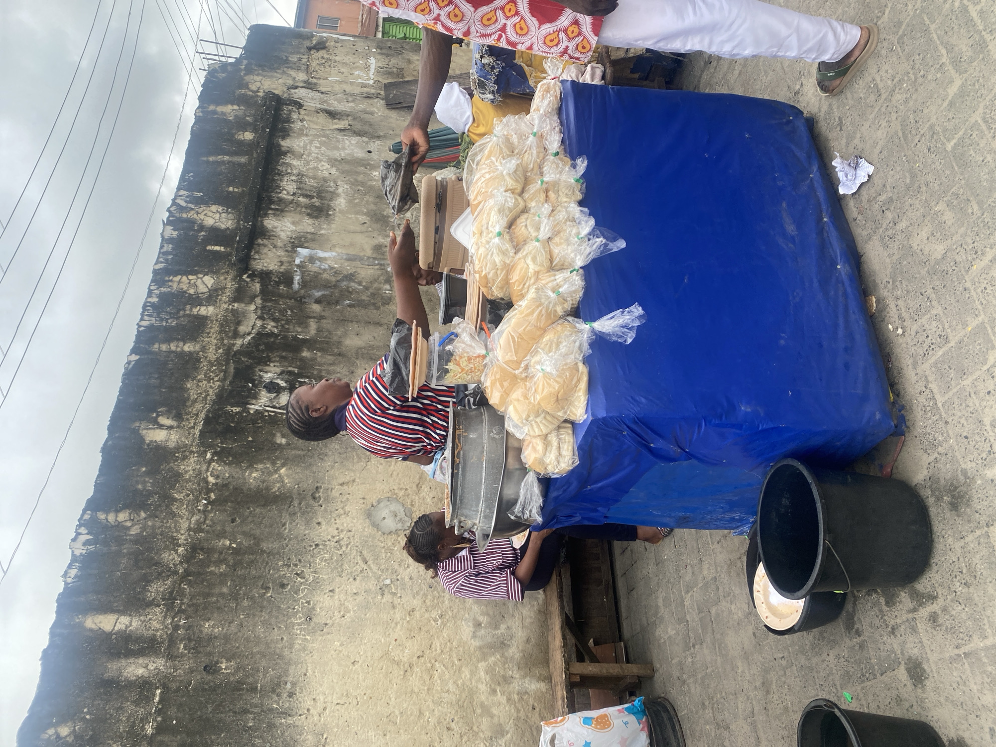 In an outdoor market setting, a vendor, a woman wearing a striped shirt, stands behind a table covered with a blue tarp, selling loaves of bread wrapped in clear plastic. A customer, wearing a white garment with a red and yellow pattern, is reaching out to receive an item from the vendor. Another woman, also in a striped shirt, sits on a wooden bench to the left, facing away from the camera, and appears to be eating. In the background, a large, weathered concrete wall dominates the scene, with some graffiti and moss growth visible. The ground is paved with grey interlocking bricks. There are several black buckets and a lighter-colored plate on the ground near the table. The sky is overcast.
