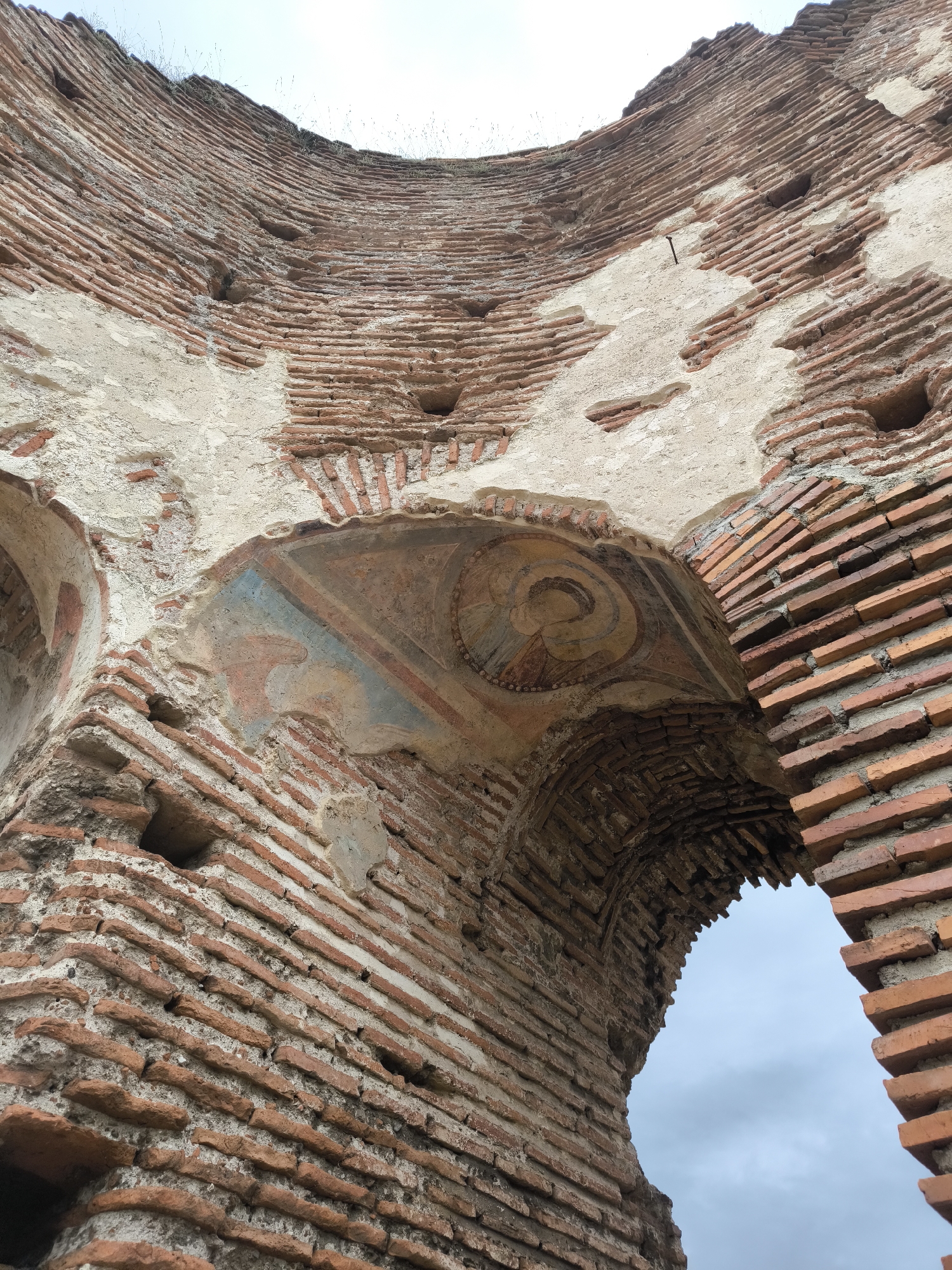 The image shows a portion of the Red Church (Chervena tsarkva) ruins, located near Perushtitsa, Bulgaria. The ruins are primarily constructed of red bricks, laid in horizontal courses with mortar. Some sections are covered with plaster or remnants of frescoes. A prominent feature is an arched opening in the brickwork, looking out to the sky. Above the arch, a section of faded fresco is visible, depicting a religious figure. The condition of the ruins is dilapidated, with bricks missing and plaster peeling. The overall impression is one of age and decay. The sky is visible through the opening.