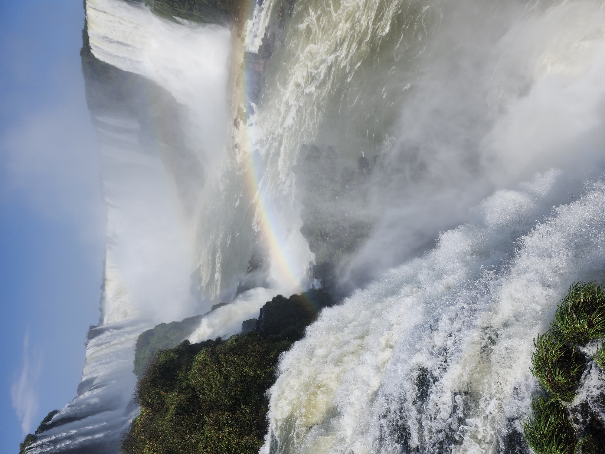 The image depicts a breathtaking panoramic view of the Iguazu Falls, a magnificent natural wonder located on the border between Argentina and Brazil, near the city of Foz do Iguaçu, Brazil. The scene is dominated by several powerful waterfalls cascading down immense cliffs, creating a dramatic and awe-inspiring spectacle. The water plunges into a turbulent river below, enveloped in a thick mist that rises high into the air.
In the mid-ground, a vibrant rainbow arches across the misty expanse, adding a touch of magic to the scene. The lush green vegetation clings to the rocky cliffs, contrasting with the white of the cascading water. The sky above is a clear, brilliant blue, providing a striking backdrop to the natural grandeur. The foreground features a close-up view of a powerful waterfall, with white water churning and crashing over the edge, and some green foliage at the very bottom. The overall impression is one of immense natural power and beauty, a testament to the raw force of nature.