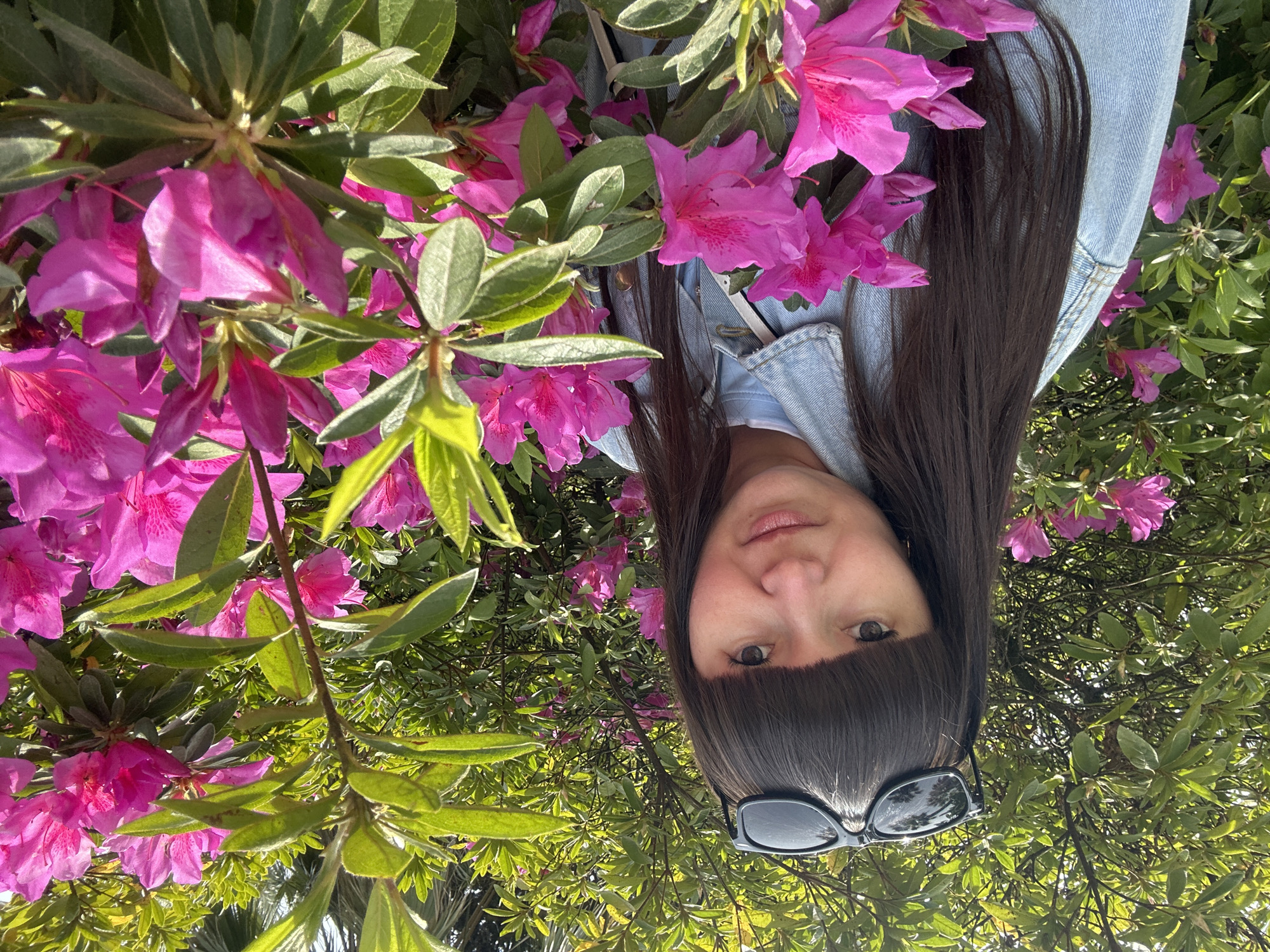 A woman with dark hair and bangs is pictured among bright pink flowers and green foliage, possibly azaleas. She is wearing a light blue denim jacket and has sunglasses perched on her head. The photo is taken from a low angle, looking up at the woman. The image is likely taken outdoors in a garden or park. There is no verifiable information to suggest the location is Sochi, Russia.