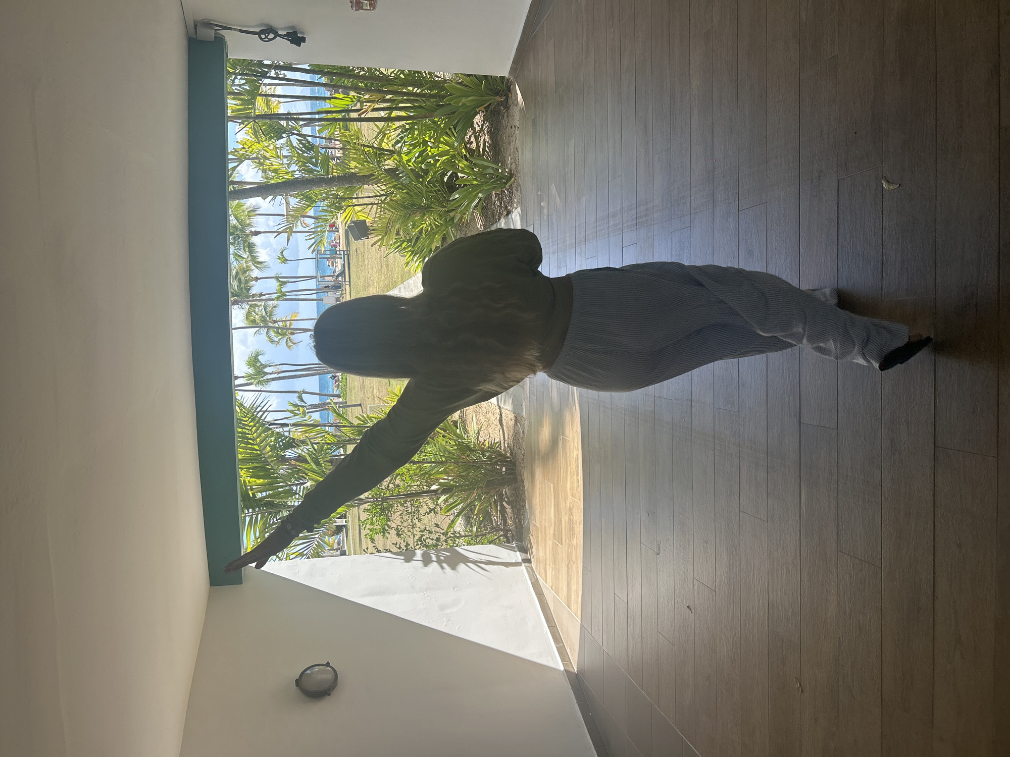 The image depicts a person, viewed from behind and partially silhouetted, standing within an open structure and facing outwards towards a tropical beach scene in Sainte-Anne, France. The individual has long, wavy light-colored hair and is wearing a dark olive-green long-sleeved top and light-colored, vertically striped wide-leg pants. Their left arm is extended upwards and outwards to the left.
The interior space features a light brown, wood-look tiled floor, white painted walls, and a white ceiling. A circular, glass-covered light fixture is mounted on the upper left wall. A turquoise-colored beam spans the top of the outdoor opening. On the upper right wall, a white electrical box with black wiring and a black, key-like object attached is visible, along with a red square device below it.
Beyond the opening, a bright exterior reveals lush green tropical vegetation, including numerous tall palm trees and various leafy plants. A manicured green lawn extends further back, leading to a sandy beach where multiple small human figures and beach furniture (umbrellas, lounge chairs) are present. The scene concludes with a wide expanse of blue ocean under a clear, bright sky. Strong sunlight from the exterior casts shadows within the indoor area.