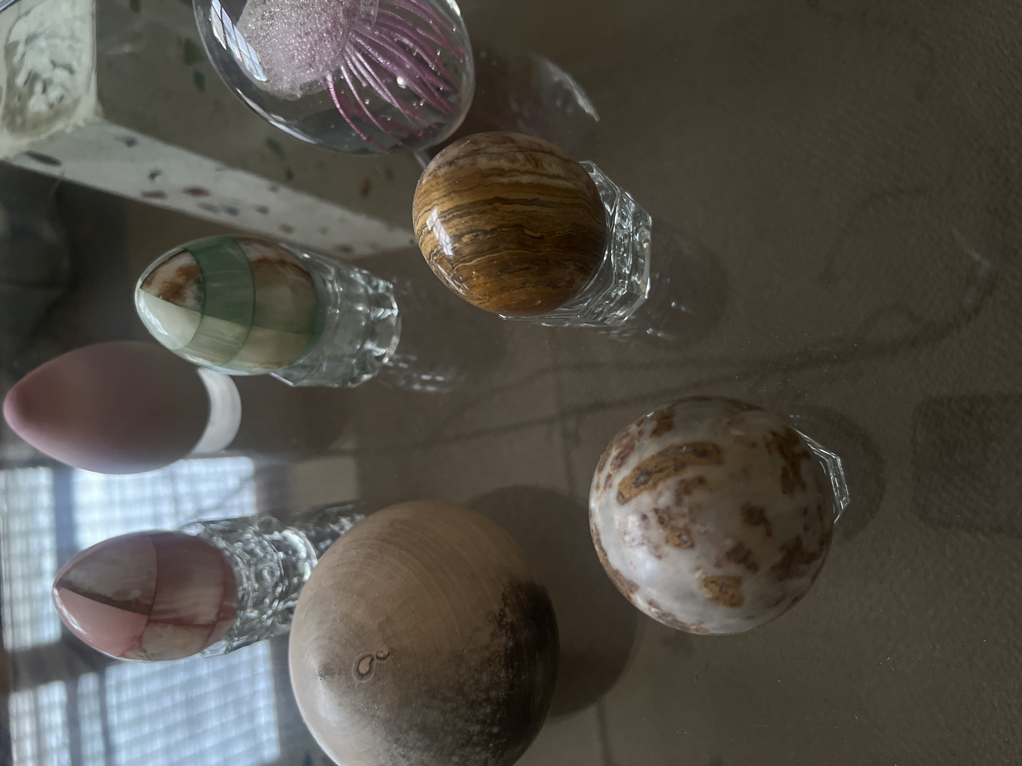 The image shows a collection of decorative glass and stone eggs resting on a reflective surface. There is a transparent glass egg filled with pink filaments. Another egg is made of a brown banded stone, resting on a small clear glass holder. A green and white striped egg sits in a glass holder. A smooth, light pink egg is positioned adjacent to a pink egg resting in a decorative glass holder. Two more eggs are visible; one made of light wood and the other a light-colored stone with brown spots. The setting appears to be indoors. The location cannot be determined.