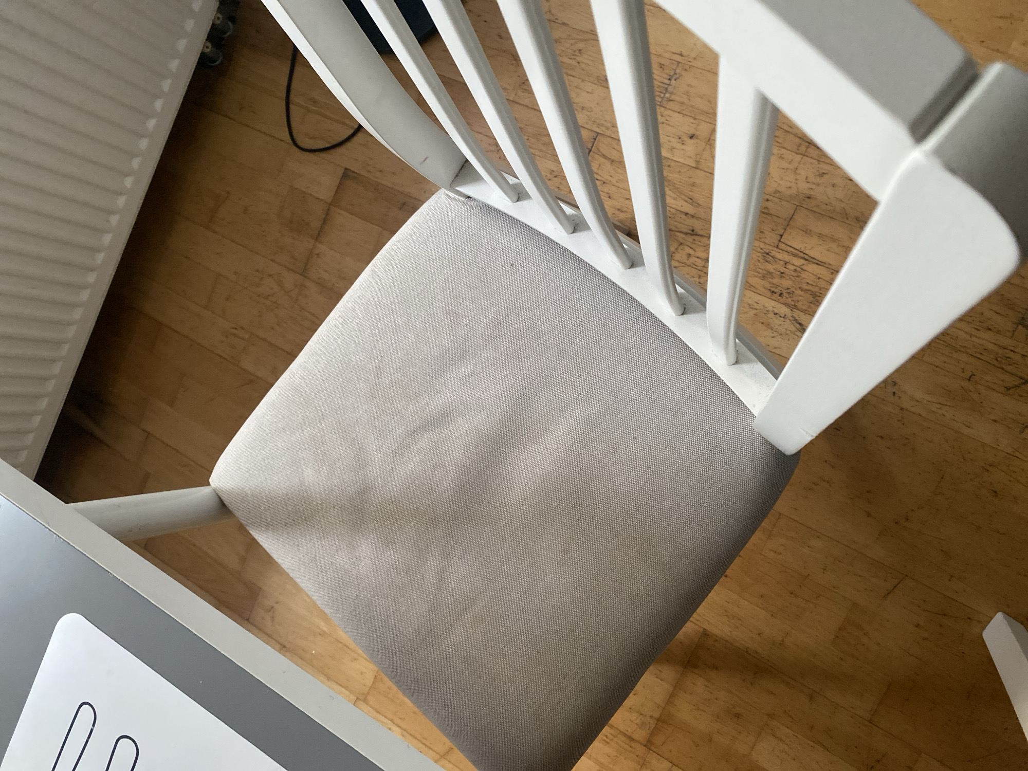 The image shows a close-up, overhead view of a grey upholstered chair with a white wooden frame, positioned on a light brown wooden floor. A white table with a grey edge is visible in the upper left corner, with what appears to be a placemat with black lines on it. A white radiator is present in the background, with a black cable lying on the floor beneath it. The scene is indoors, with no specific identifying features to pinpoint a location within Wrocław, Poland.