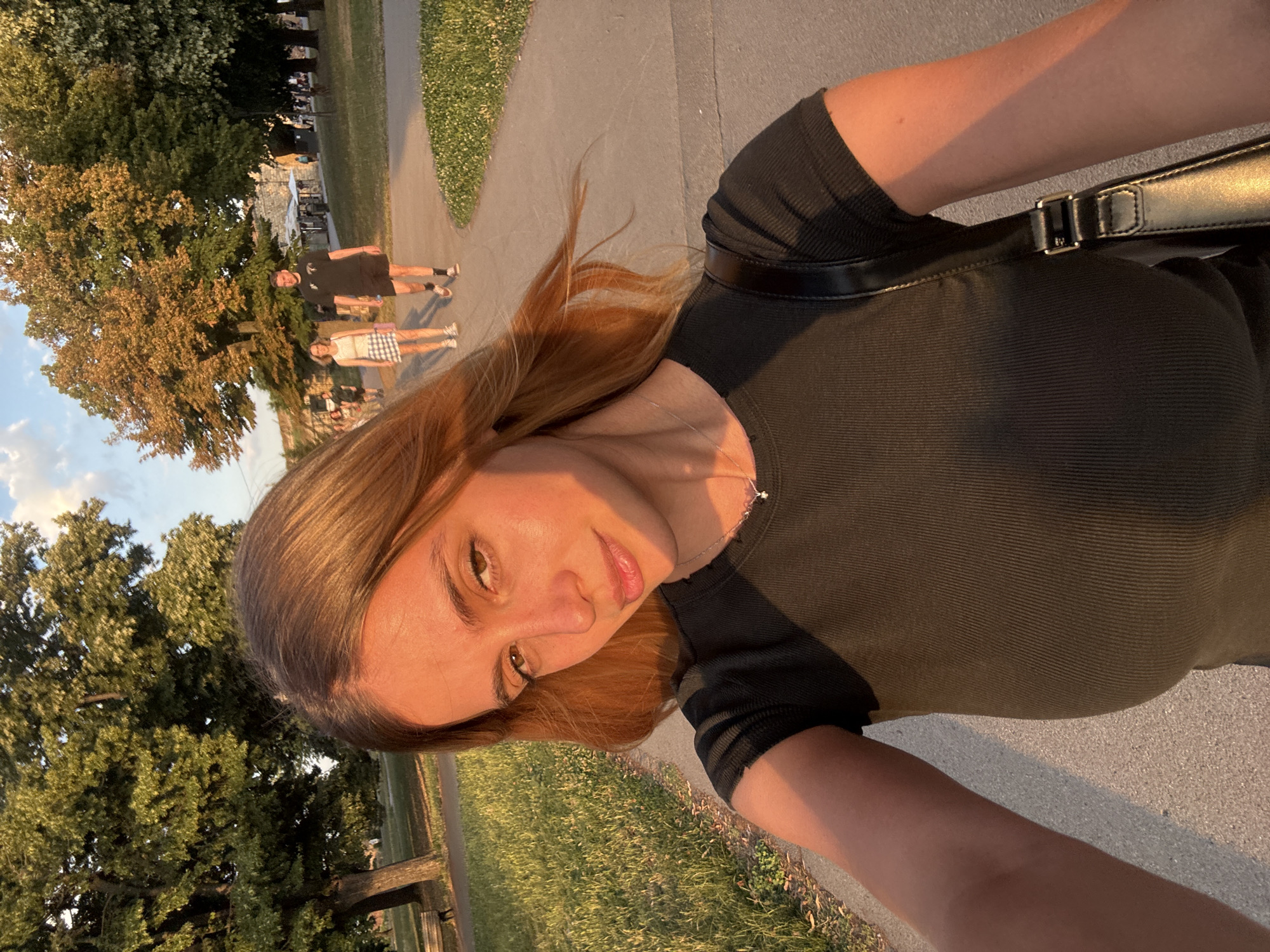 A young woman is taking a selfie outdoors on a paved path with grass and trees lining the sides. She is wearing a dark, ribbed t-shirt and a silver necklace. A black purse with a silver buckle is slung over her shoulder. In the background, a man and a woman are walking away from the camera. The man is wearing a black t-shirt and shorts, while the woman is wearing a white top and a plaid skirt. The scene appears to be a park, possibly in Belgrade, Serbia, given the architectural style of the buildings visible in the distance and the types of trees. The lighting suggests it is late afternoon or early evening, with the sun casting long shadows and highlighting the golden tones of the woman's hair.