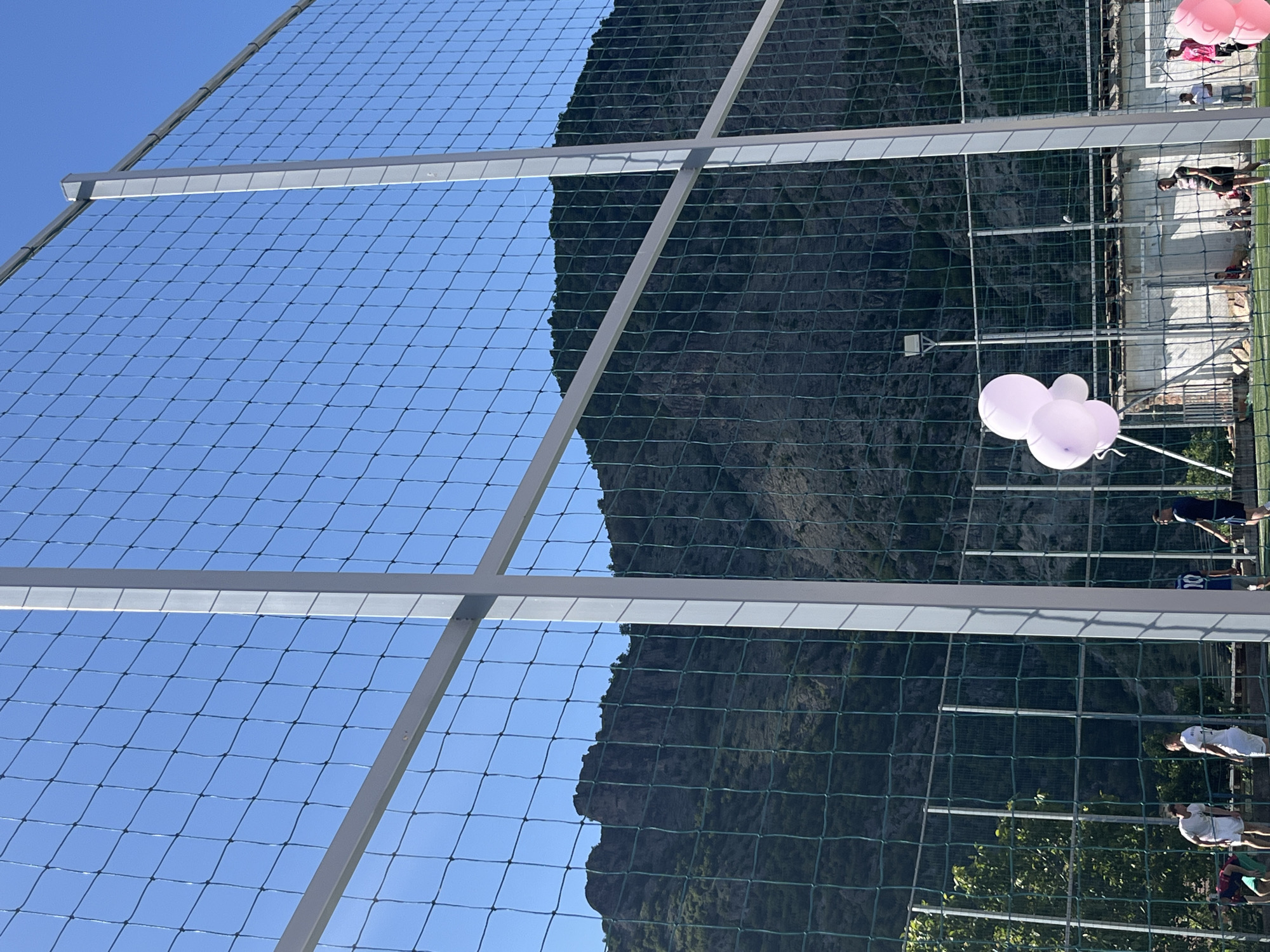 The image shows a mountain backdrop viewed through a net. A white metal frame supports the net. The sky is blue. Pink and white balloons float near the net. People stand near a wall on the right side of the image. The location is Bukovo, Bulgaria.