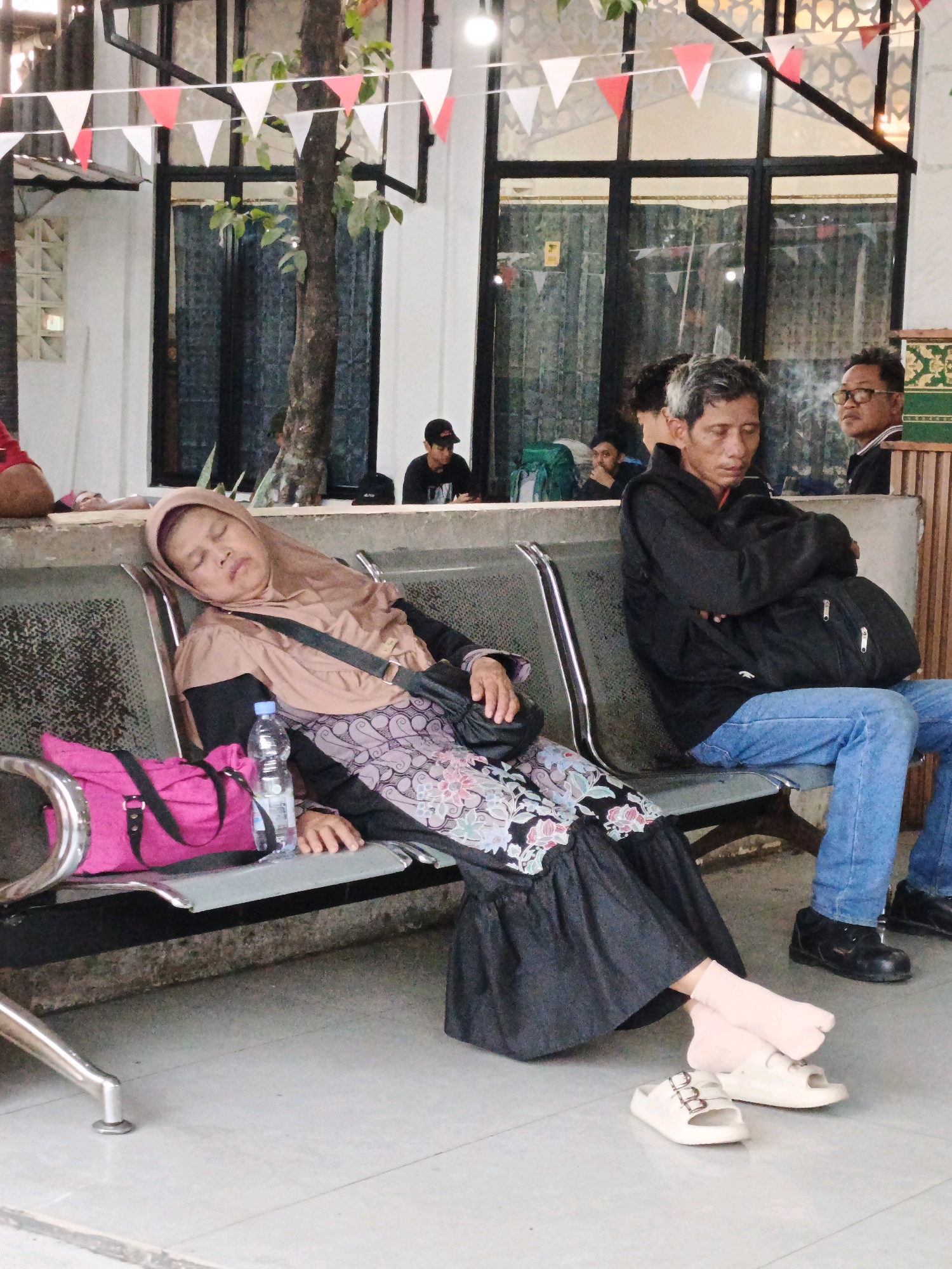 An elderly woman wearing a hijab and a long dress is sleeping on a metal bench. Next to her is a pink bag and a bottle of water. To her right, a man in a black jacket and blue jeans is also sitting on the bench, with his arms crossed. In the background, other people are visible, some seated and some standing. The scene appears to be an outdoor waiting area, possibly at a bus station or a public gathering place in North Jakarta, Indonesia, with decorative flags strung overhead. The environment is somewhat cluttered with some trees and architectural elements visible.