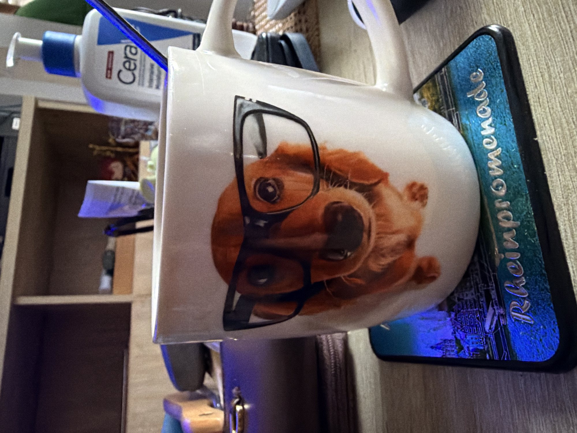 This is a close-up, eye-level, indoor shot of a white mug with a graphic of a dachshund wearing glasses. The mug is placed on a coaster depicting a European cityscape with the text "Rheinpromenade" on it. In the background, a bottle of CeraVe moisturizer is visible on a shelf. The scene appears to be on a desk or table, with other objects and furniture subtly blurred in the background. There is no indication of the location being Castillo Bajo-conejito, Spain.