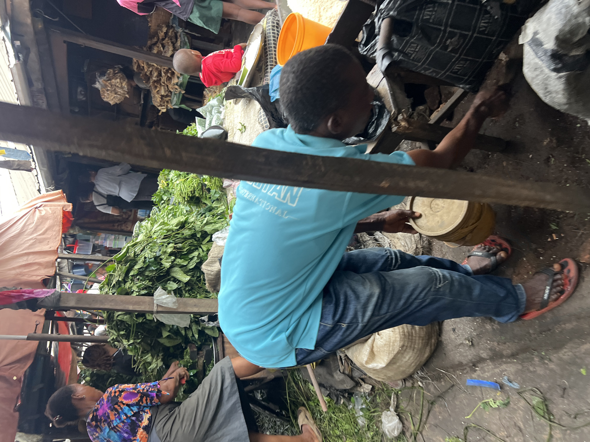 In Nkpor, Nigeria, a bustling market scene unfolds. A woman in a colorful blouse and dark skirt is seen preparing vegetables on a wooden surface. Beside her, a large mound of fresh green produce dominates the scene. In the foreground, a man wearing a light blue t-shirt and distressed denim jeans is actively engaged, bending down to sift through what appears to be soil or debris. He holds a light-colored bucket in his left hand. The ground is a mix of dirt and scattered leaves, with various sacks and bags visible around the marketplace. A small child in a red hoodie observes the activities from a distance. The overall environment suggests a typical day at a vibrant Nigerian open-air market.