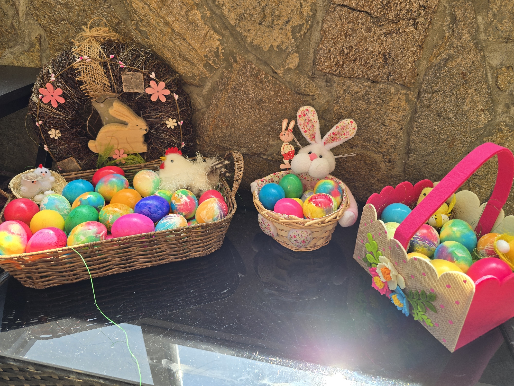 Three Easter baskets and a wreath rest on a black, reflective surface. The surface is outdoors, with a rough, tan stone wall forming the backdrop. The leftmost basket is a large, rectangular woven basket overflowing with brightly colored Easter eggs, a small toy chicken in a nest, and a larger white stuffed chicken. Next to it is a smaller woven basket filled with Easter eggs and a white stuffed bunny with flowered ears, next to a small, wooden bunny figure. The third basket, to the right, is a pink and beige felt basket with a pink handle, also filled with Easter eggs and small toy chicks. Behind the left basket is a wreath made of twigs, with a wooden bunny and flowers attached. A thin, green string lies on the surface in front of the left basket. Burgas, Bulgaria, is not present.
