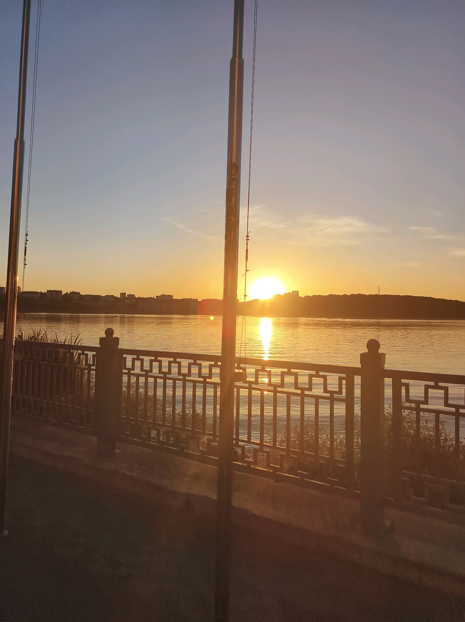 This image captures a sunset over the Ternopil Pond in Ternopil, Ukraine. The sun is low on the horizon, casting a warm, golden light across the water and illuminating the sky with hues of orange and blue. In the foreground, a decorative metal railing lines the promenade, separating it from the pond. Beyond the railing, reeds and vegetation grow along the water's edge. The far bank of the pond features a cityscape with buildings and trees silhouetted against the bright sky. Two tall, slender poles, possibly flagpoles, stand prominently on the left and right sides of the frame, their metallic surfaces reflecting the sunset light. The overall scene depicts a serene urban waterfront at dusk.