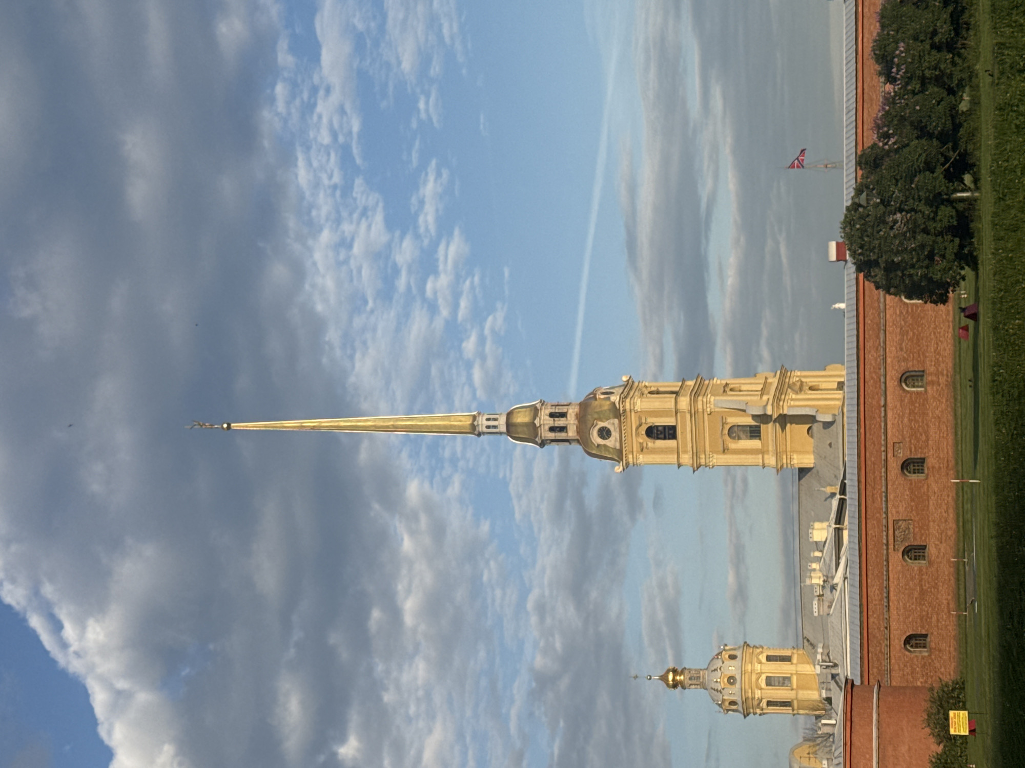 This image captures a majestic view of the Peter and Paul Fortress in Saint Petersburg, Russia, under a dynamic sky.
The most prominent feature is the towering golden spire of the Peter and Paul Cathedral, positioned centrally. It culminates in a distinctive golden angel or weather vane. Below the spire, the ornate golden dome and detailed architectural elements of the cathedral's upper sections are visible, showcasing intricate design and craftsmanship. To the lower right of the main spire, a smaller golden-domed structure, possibly another part of the cathedral or an adjacent building within the fortress complex, can be seen.
In the lower portion of the image, a robust red brick building, indicative of the fortress walls, runs across the frame. It features multiple arched windows set into its facade and is bordered by lush green grass and some trees.
The sky above is a blend of clear blue patches and various cloud formations, including wispy white clouds and more substantial grey clouds. A faint airplane contrail is visible streaking across the upper sky. The lighting suggests either early morning or late afternoon, as the golden structures are brightly illuminated, contrasting with clear shadows. In the distance, a flag, which appears to be the Saint Andrew's flag (Russian Navy flag), is flying above a remote structure.
The scene conveys a sense of historical grandeur and architectural beauty. There are no people visible, and no discernible text can be read in the image.