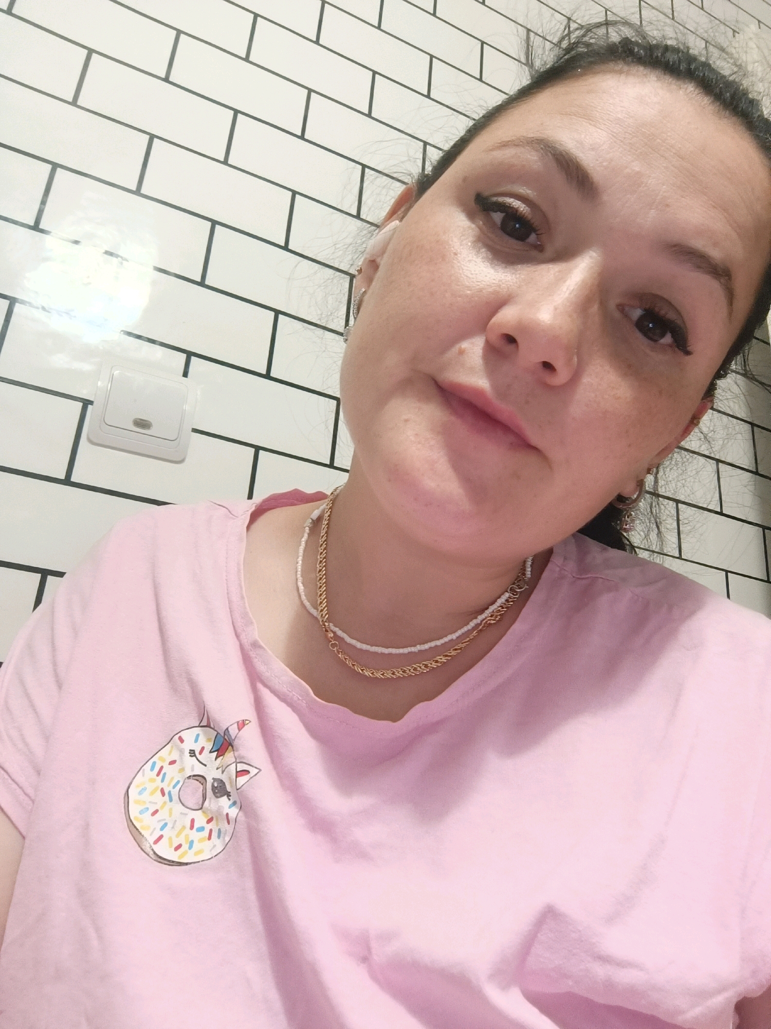 A medium shot captures a woman with dark hair pulled back, wearing a light pink t-shirt with a cartoon donut graphic. She has freckles across her face and is wearing subtle makeup. Two necklaces adorn her neck: a delicate white beaded chain layered over a thicker gold rope chain. She has a small hoop earring in her left ear. The background is a white subway tile wall with dark grout lines, and a white light switch is visible on the left side. The scene appears to be indoors, possibly a bathroom or kitchen. There is no indication of the city of Chaikovsky, Russia.