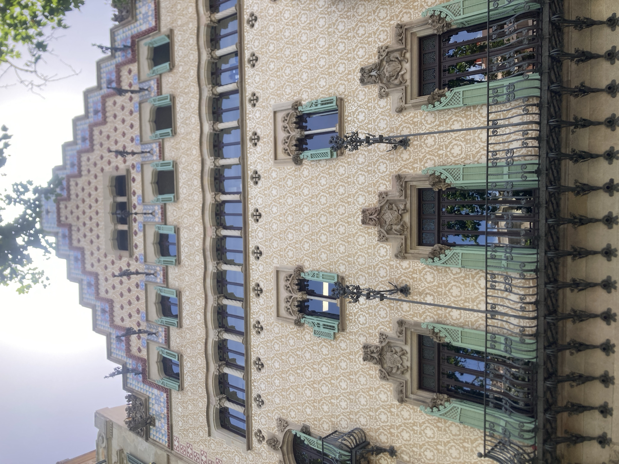 This is a low-angle exterior, eye-level shot of the Casa Batlló in Barcelona, Spain. The building's facade is decorated with a mosaic of colorful tiles and features a distinctive roofline with ornate chimneys. The main body of the building is covered in a repeating pattern of organic motifs. Several windows with green shutters are visible, as well as wrought-iron balconies. The sky is bright and sunny, with some tree branches visible at the top of the frame. No people are visible in the image.