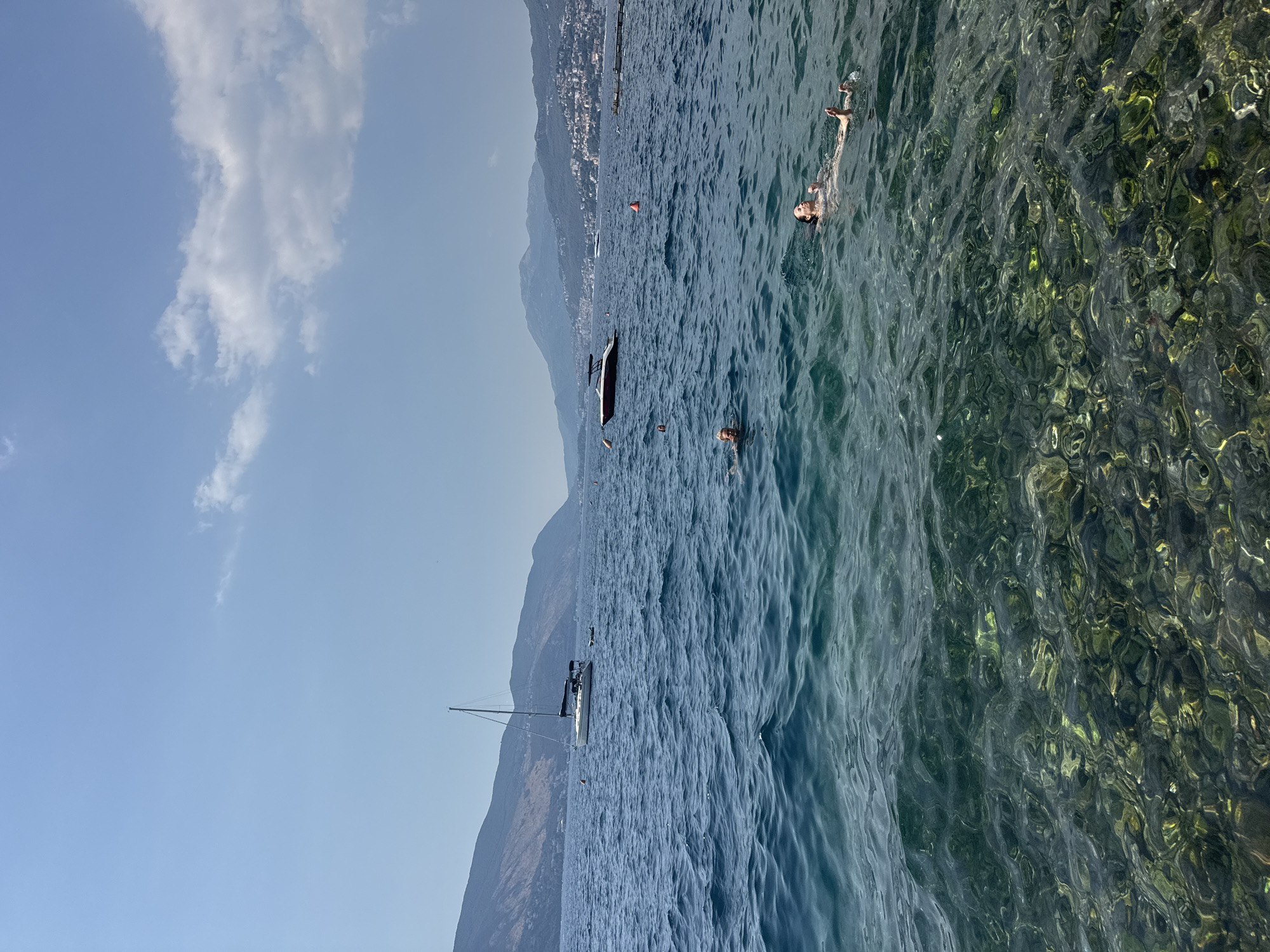 In a wide shot of a calm, sun-drenched bay, several people are swimming in the clear, turquoise water. The foreground features a rocky seabed visible through the translucent water. In the middle ground, two individuals are swimming, their heads bobbing above the surface. One person is floating on their back, arms outstretched, while another is treading water. Further out, a white sailboat with a tall mast is anchored, with several people on its deck. A small, dark-colored motorboat is also visible in the distance. The background is dominated by rolling, tree-covered hills that recede into the hazy horizon. The sky is a clear, pale blue with a few wispy white clouds. The scene is tranquil, evoking a sense of summer leisure.