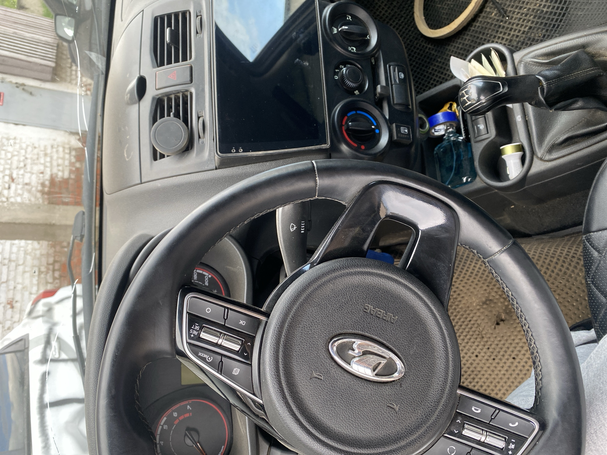 The image depicts the interior of a vehicle, specifically the dashboard and steering wheel. The steering wheel has the Lada logo, suggesting it is a Lada vehicle. The dashboard features controls for climate and an infotainment system. The gear shift is visible with some small items in the cup holders. The scene appears to be in Chaikovsky, Russia, due to the Lada branding and location.