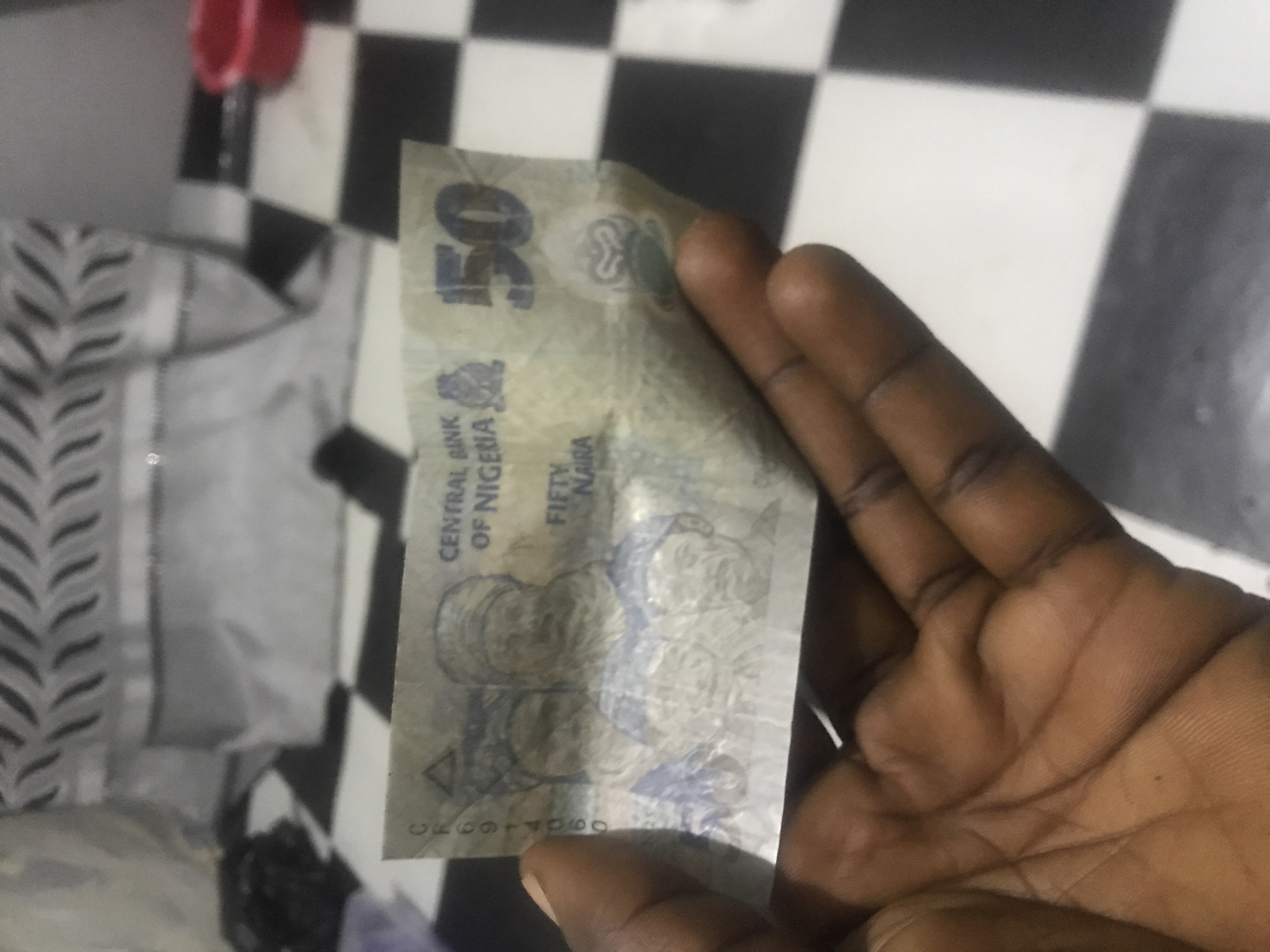 A close-up, slightly rotated image shows a dark-skinned hand holding a worn Nigerian 50 Naira banknote. The banknote is held vertically, with its front face visible. Prominent text on the banknote includes a large "50" at the top left, "CENTRAL BANK OF NIGERIA," and "FIFTY NAIRA" written below. A serial number, "C399171990," is visible at the bottom left. The banknote also features indistinct portraits of two individuals.
The background is mostly composed of a black and white checkered pattern, likely a tiled floor. To the left, an out-of-focus grey garment with a black chevron or zigzag pattern is visible. A small, blurry red object can be seen in the top left corner. The scene appears to be indoors in Rafin Par, Nigeria, with a bright, even light source. No specific activity beyond holding the currency is depicted.