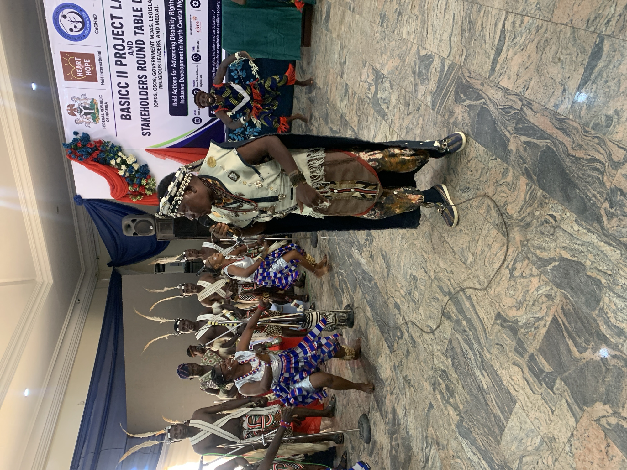 A group of men and women in traditional attire, some wearing elaborate headwear with feathers, are performing a dance in front of a banner. The banner reads "BASIC II PROJECT LAUNCH AND STAKEHOLDERS ROUND TABLE". A man in the foreground is singing into a microphone, dressed in a distinctive outfit with intricate beadwork and patterned legwear. The floor is tiled with a marbled pattern, and several microphones on stands are positioned around the performers. The event appears to be an indoor gathering, possibly a cultural festival or a formal presentation. The location is stated to be in the city of Gura Top, Nigeria.