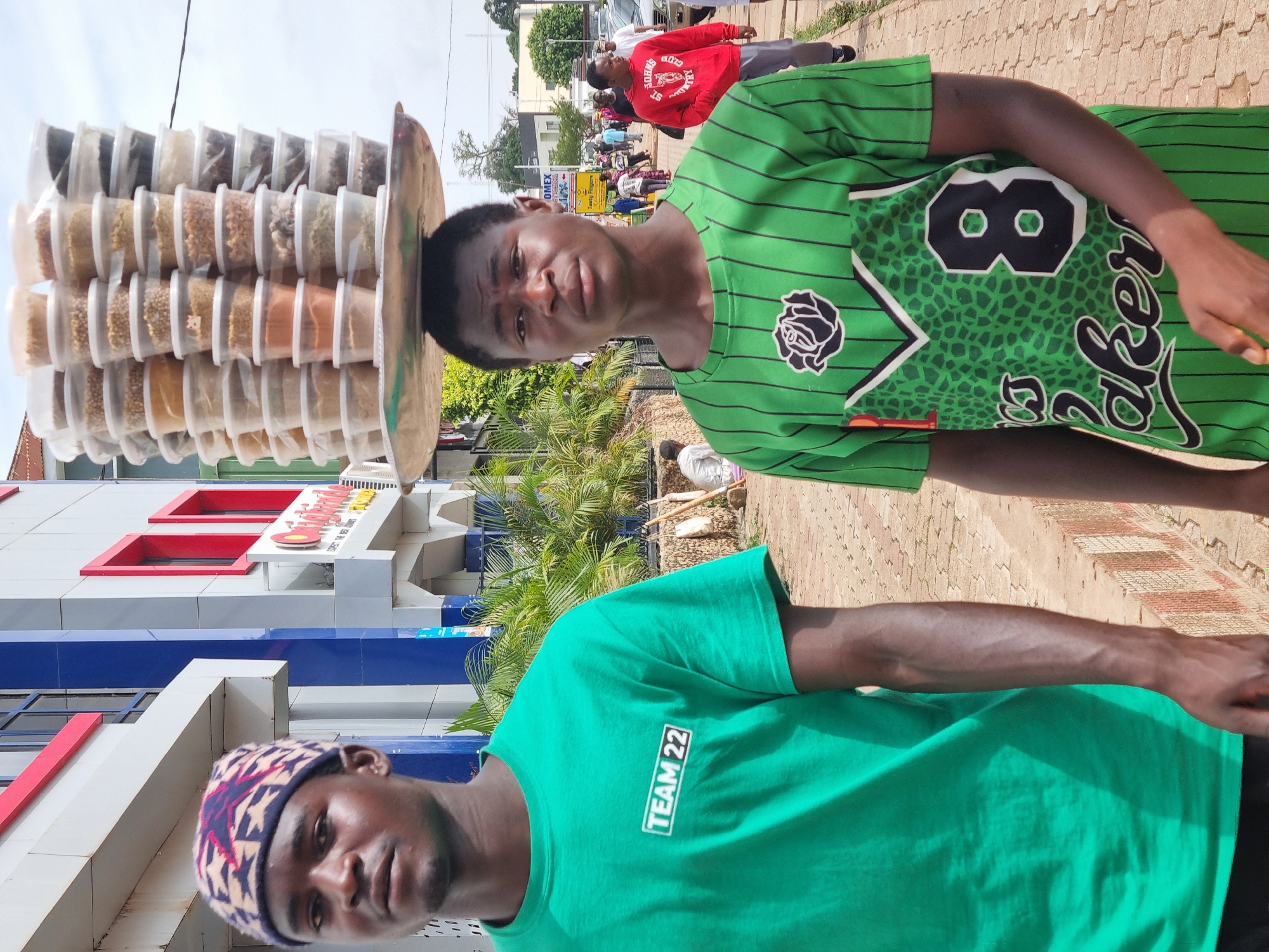 Two young men stand on a brick sidewalk in what appears to be a commercial area of Jos, Nigeria. One man wears a green t-shirt and a patterned skullcap. The other man, further away in the background, wears a green striped jersey and balances a wide, flat tray piled high with small containers filled with assorted spices or food items on his head. A building with blue and white horizontal stripes and red window frames stands to the left. A pedestrian in a red shirt walks on the sidewalk behind the men. Other pedestrians and vehicles are visible in the background, suggesting a busy street scene.