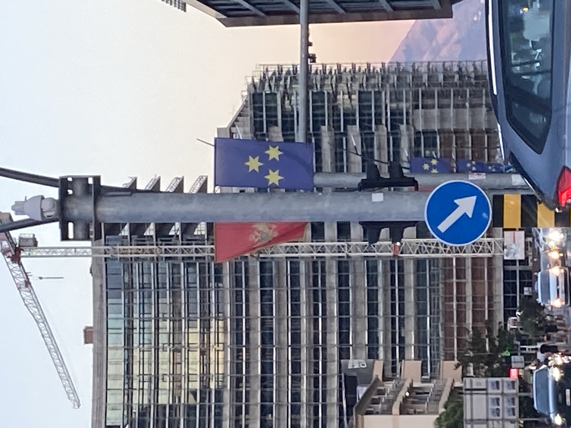 The image shows a street scene in Budva, Montenegro. A tall, modern building under construction, with a crane visible, dominates the background. In the foreground, a traffic light pole displays two flags: one red with a gold double-headed eagle and crown (Montenegro), and another blue with three yellow stars (European Union). A blue circular traffic sign with a white arrow pointing down and to the left is also visible on the pole. Cars are driving on the street, and several pedestrians are walking on the sidewalk. The sky is overcast with a hint of pink from sunset.