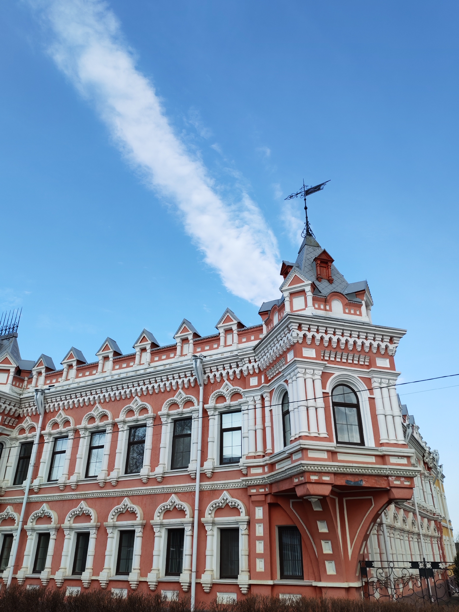 The image features a detailed view of the facade of the Popov House, a historical building located in Sarapul, Russia. The architecture displays intricate ornamentation in a combination of salmon and white hues. The building's design incorporates a protruding corner section with rounded windows, and an elaborate roofline that includes small towers and a weather vane. A bright blue sky with a single, long white cloud serves as a background. Bare shrubbery is visible at the base of the building. The lighting suggests it is daytime.