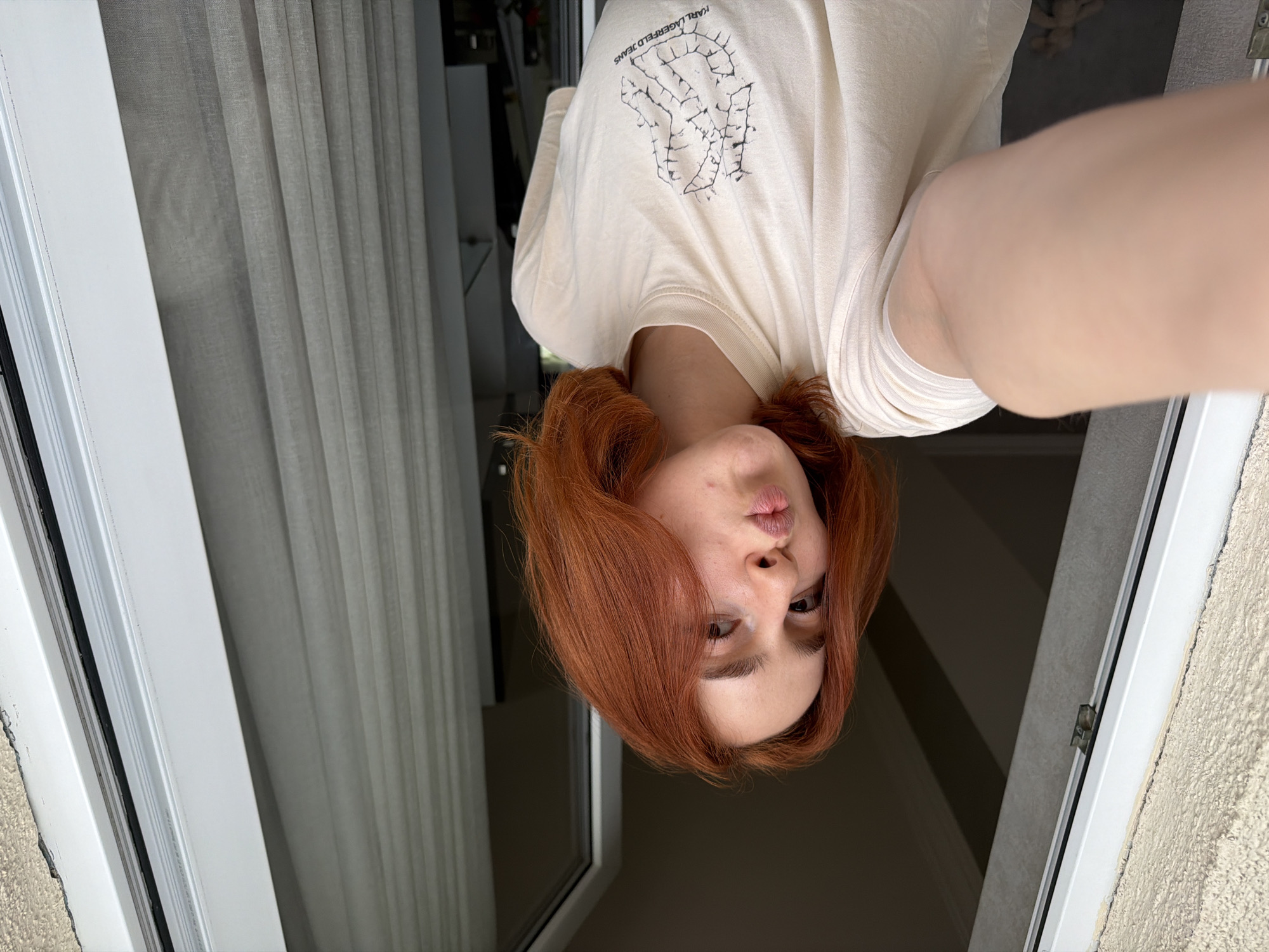 A selfie, taken at an approximate 180-degree angle, presents a woman with red hair and fair skin. She wears a light-colored T-shirt with black graphics. The environment suggests an interior with a window or door frame on either side of her. Curtains hang on the left. Outside the door/window on the right is stucco wall, likely exterior. There are no discernible landmarks to indicate the location is Tbilisi, Georgia.