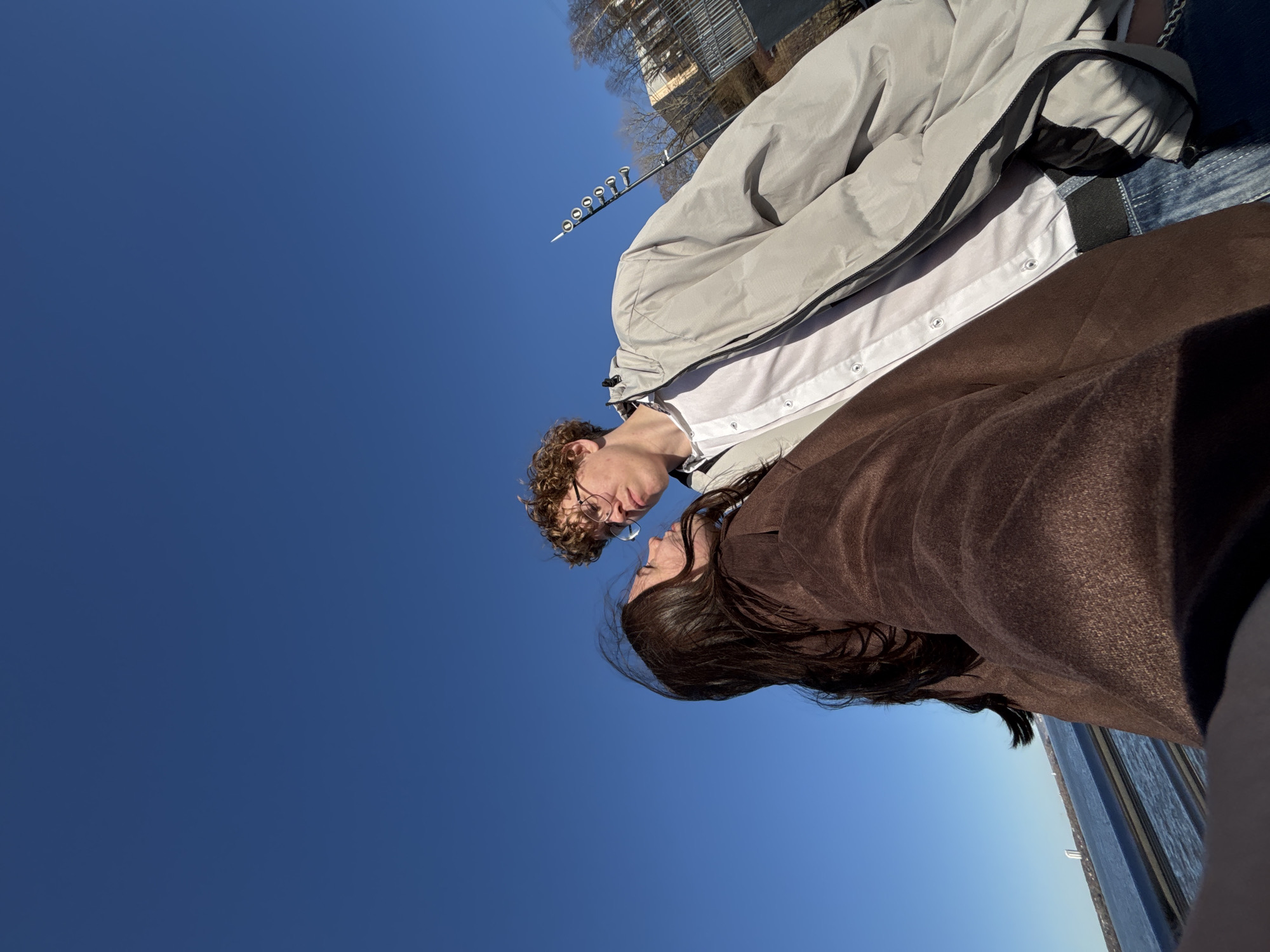 A young couple are face-to-face, likely on a bridge, on a clear day with a bright blue sky. The person on the left has curly blonde hair and wears glasses, a white collared shirt, and a light gray jacket. The person on the right has long, dark hair and wears a brown jacket. In the background, there is a building and a row of metal streetlights, suggesting an urban location, probably in Perm, Russia. The photo appears to be taken at an angle looking up at the couple.