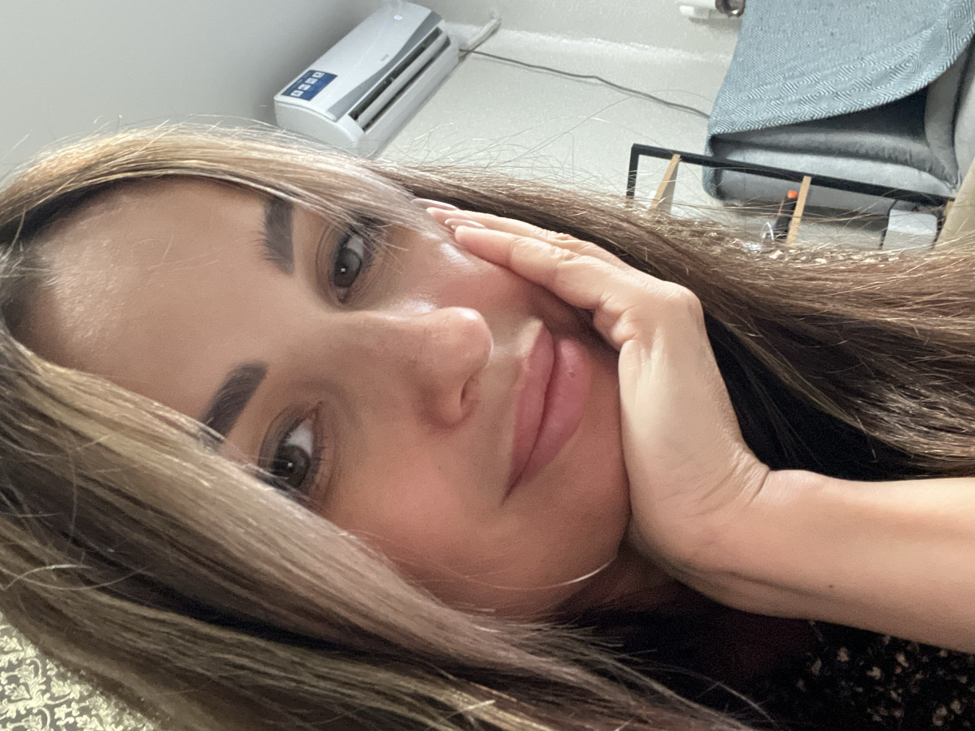 A close-up, waist-up, indoor shot shows a woman from the chest up, with her left hand propping up her chin and her right hand near her cheek. Her head is tilted to the right and her eyes are looking at the camera. She has long, light brown hair that is partially covering her face. Her lips are slightly parted and a subtle smile is on her face. The background includes a white air conditioning unit on a light-colored wall, a shelf with some objects on it, and a blue patterned blanket draped over furniture. The lighting is soft and appears to be natural.