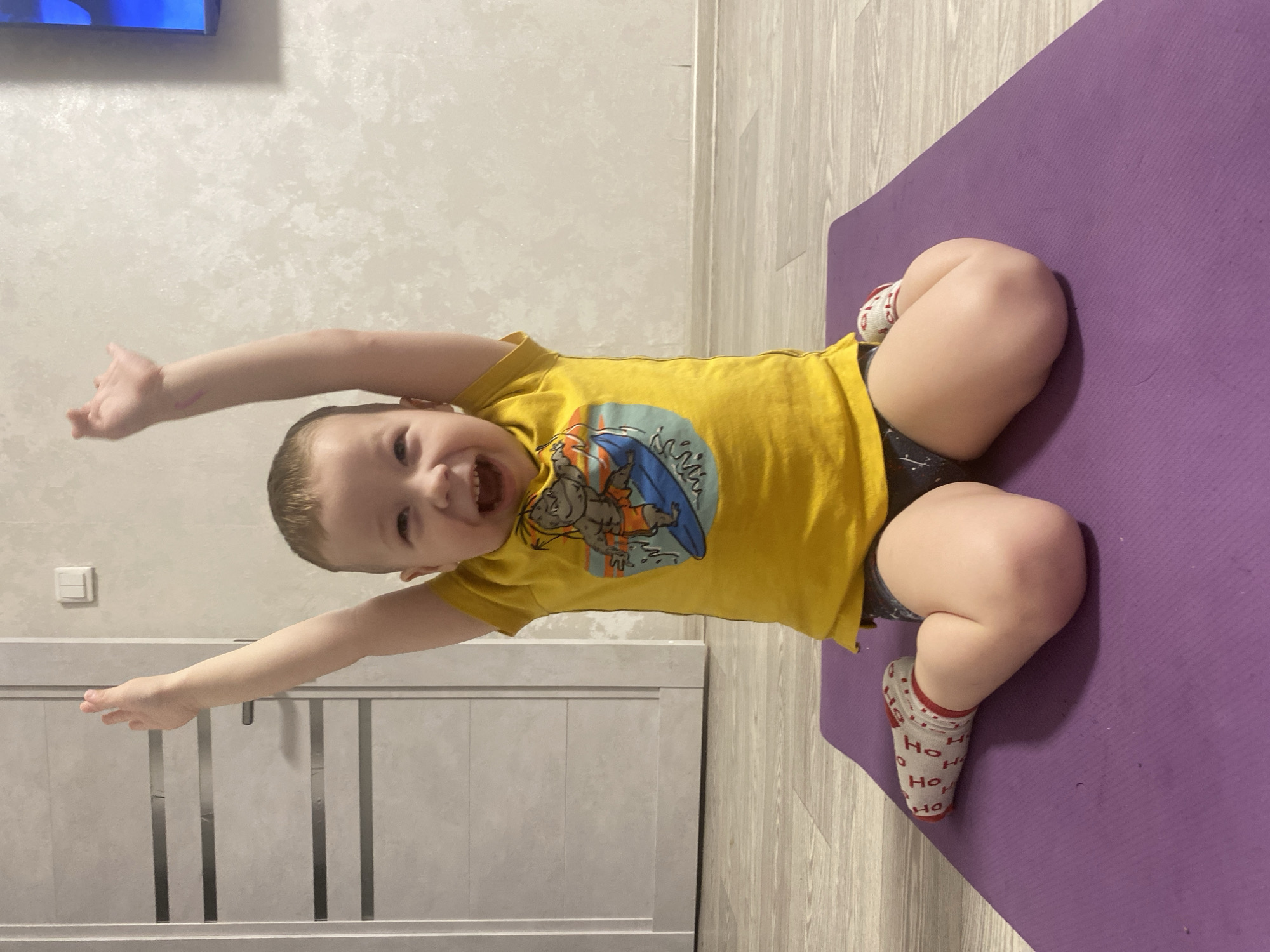 A young child is lying on a purple yoga mat on a light-colored wood floor. The child is wearing a yellow t-shirt with a surfer graphic and dark-colored shorts. The child is also wearing white socks with red "HO" patterns. The child is smiling and has their arms outstretched. The background includes a light-colored wall and a light-colored cabinet. The location is not specified to be Sochi, Russia, in the image.