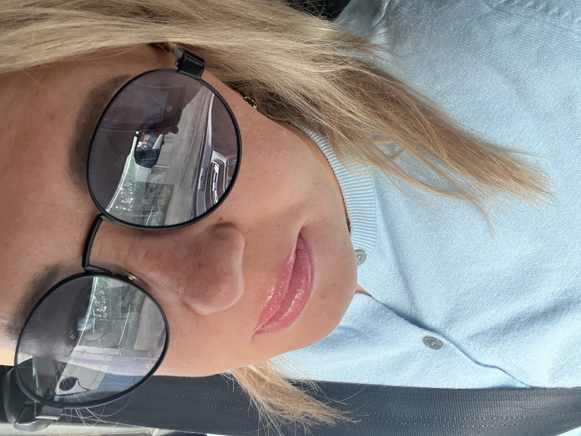 A close-up, outdoor, daylight, eye-level, waist-up selfie shows a blonde woman in sunglasses and a light blue sweater. The woman is in the driver's seat of a vehicle, indicated by the car's interior reflected in her sunglasses and the seatbelt visible. The background visible through the sunglasses appears to be a street scene in a city, possibly with buildings and trees. The woman's face is partially obscured by her sunglasses, but her lips are visible and appear to be wearing lip gloss. The overall lighting suggests it is daytime. No specific landmarks or details indicate the location is Chaikovsky, Russia.