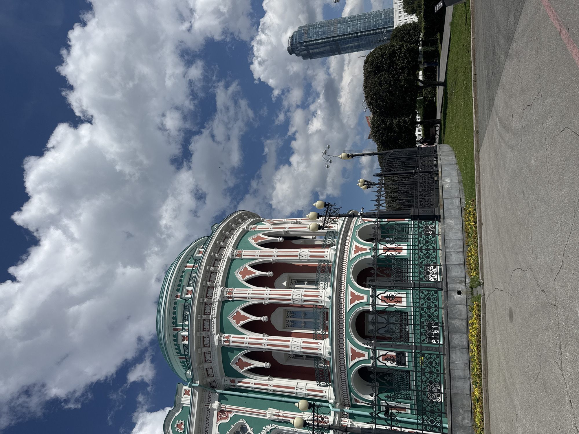 The image captures a daytime exterior scene in Yekaterinburg, Russia, featuring a prominent, ornately decorated building and a modern skyscraper in the background. The foreground shows a paved area with cracked asphalt, leading to a low, manicured garden bed with yellow flowers. A black wrought-iron fence separates the garden from the sidewalk and the building's grounds.
The building itself is a striking structure with a curved facade. It is painted in vibrant teal and red, with white decorative accents around the arched windows and columns. The architectural style appears to be a mix of classic and ornate elements, with multiple levels of balconies and arched openings. Several black lampposts with multiple globes are visible along the sidewalk in front of the fence.
In the middle ground, a patch of green lawn is visible, with two well-maintained, rounded trees providing shade. A park bench is placed beneath one of the trees. Two individuals are casually walking in the grassy area.
Dominating the background on the right is a very tall, modern glass skyscraper, distinctive for its tiered, rounded design. Its blue-tinted glass reflects the sky and clouds. Other smaller buildings are visible in the distance, suggesting an urban setting. The sky is a deep blue, dotted with large, fluffy white clouds. The overall impression is one of a city with a blend of historical and contemporary architecture under a bright, partly cloudy sky. The location is identified as the Yekaterinburg City Hall, also known as the Plotinka embankment.