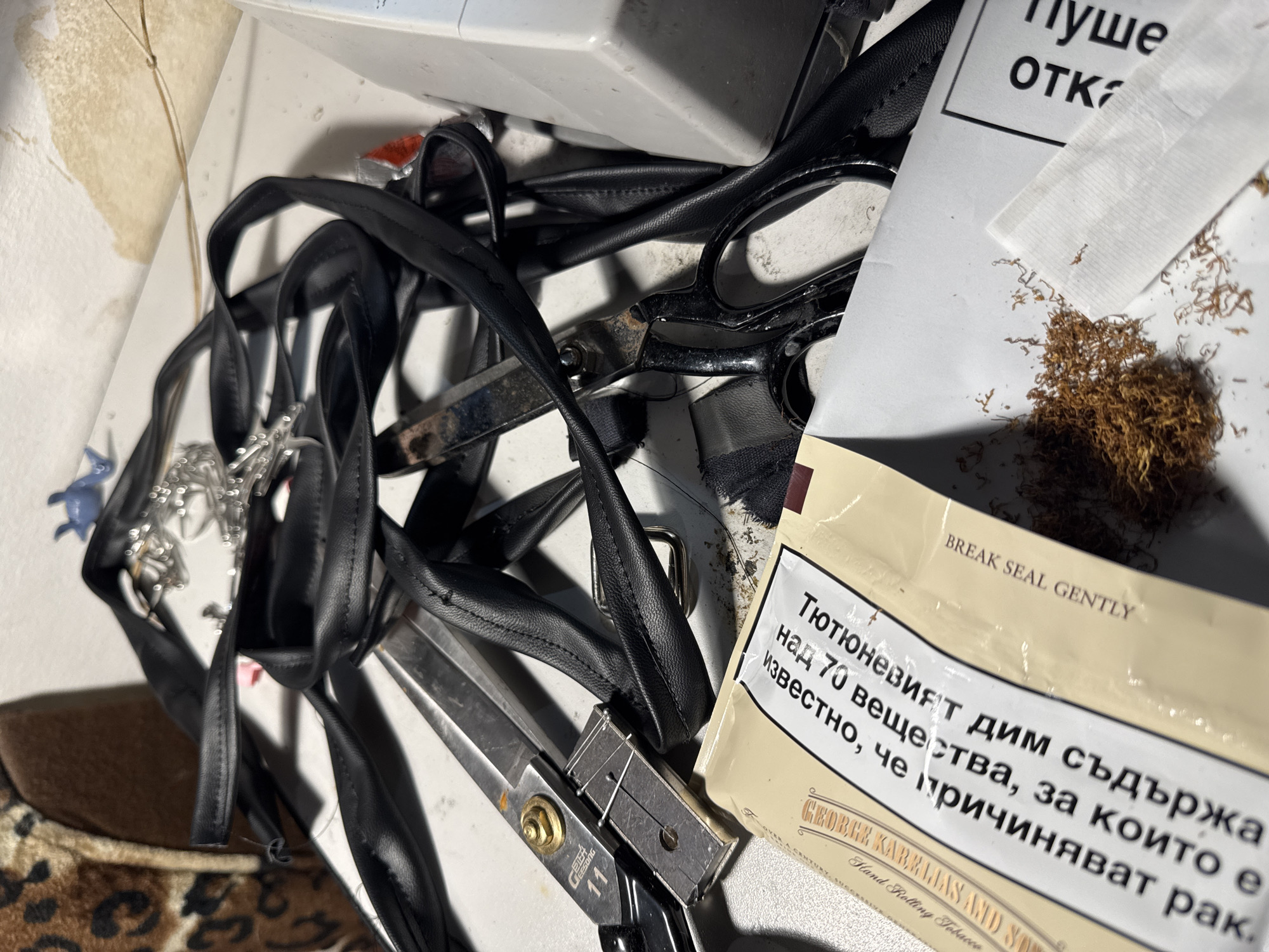 The image presents a cluttered tabletop scene featuring crafting or repair-related items. A mass of black leather straps is entangled with two pairs of scissors: one pair appears smaller and silver-colored, while the other is larger with black handles. Adjacent to these items, there's a packet of rolling tobacco, labeled "George Karelias and Sons," along with loose tobacco strands and a rolling paper. Text on the packet is in Bulgarian, indicating the product contains "Тютюневият дим съдържа над 70 вещества, за които е известно, че причиняват рак" (Tobacco smoke contains over 70 substances that are known to cause cancer). A label with "Пуше ОТК" is also present. The items are spread on a white surface with signs of wear, including what appear to be wallpaper edges, suggesting a domestic setting.
Based on the Bulgarian text on the tobacco packaging, it is likely located in Bulgaria, and without further information, it is reasonable to suggest a city with commerce and tobacco use, such as Sofia.