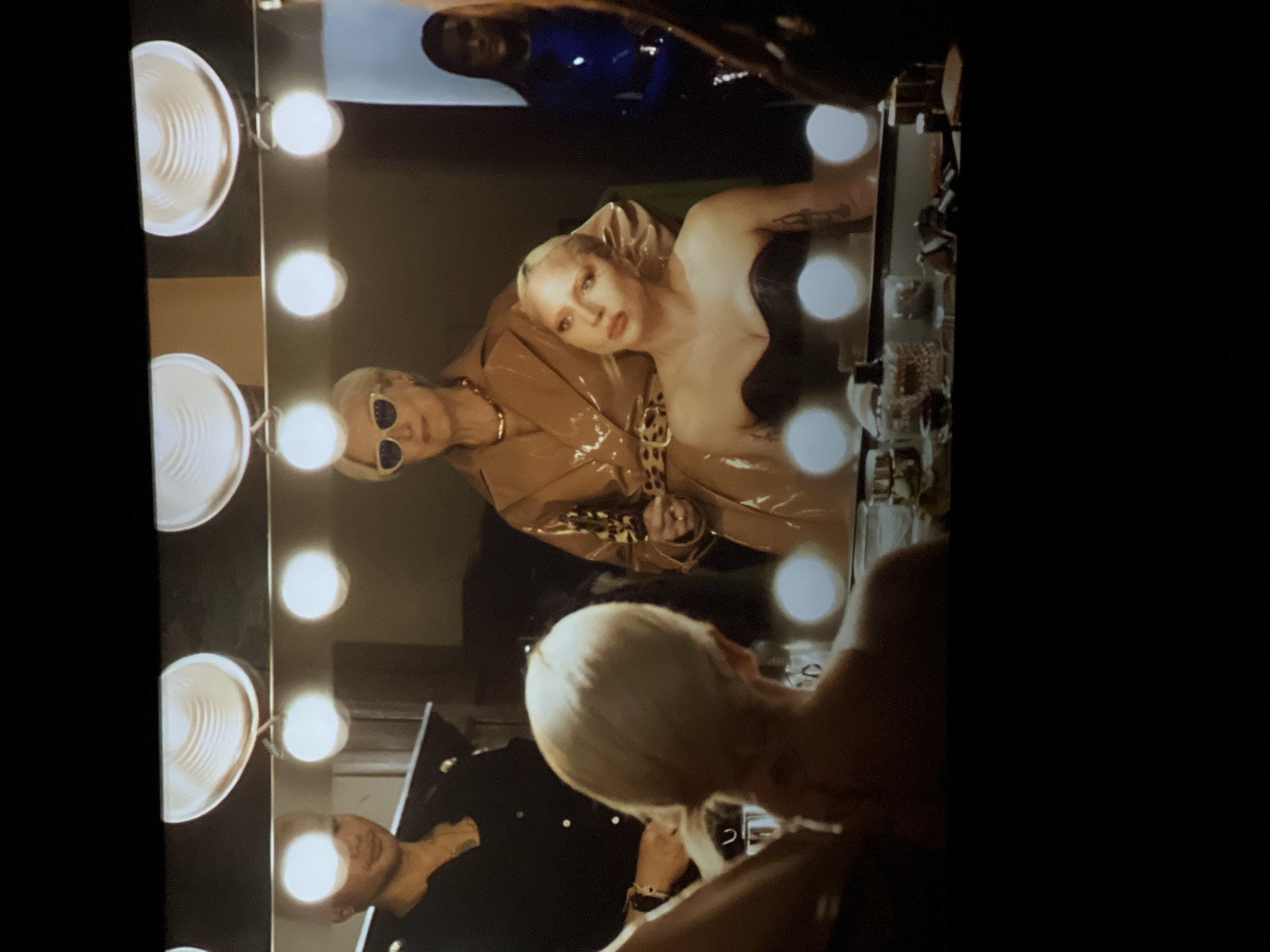The image shows a makeup mirror with multiple lights surrounding its perimeter. Reflected in the mirror is a woman with blonde hair, wearing a black top and a tan jacket with leopard print accents. Another person with short blonde hair and sunglasses is partially visible beside her. In the background, a dark-skinned person is wearing a shiny blue outfit. In the foreground, a person with blonde hair is seated, possibly receiving makeup or hair styling. Another person in a black shirt is standing behind this seated person. The location is not discernable based on the image, therefore I cannot assert that it is in Yerevan, Armenia.