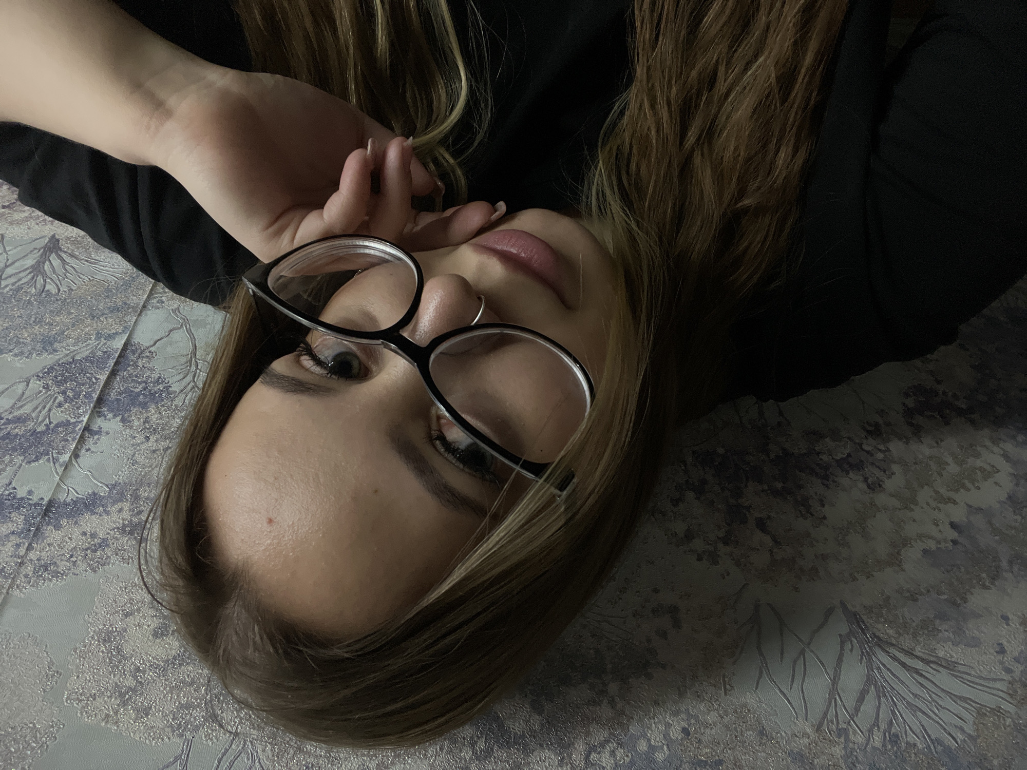 A woman lies on a patterned surface, possibly a bedspread. She wears glasses and a dark-colored top. Her face is oriented towards the camera. Her hair is light brown. Her hand is near her face. No location information can be verified.