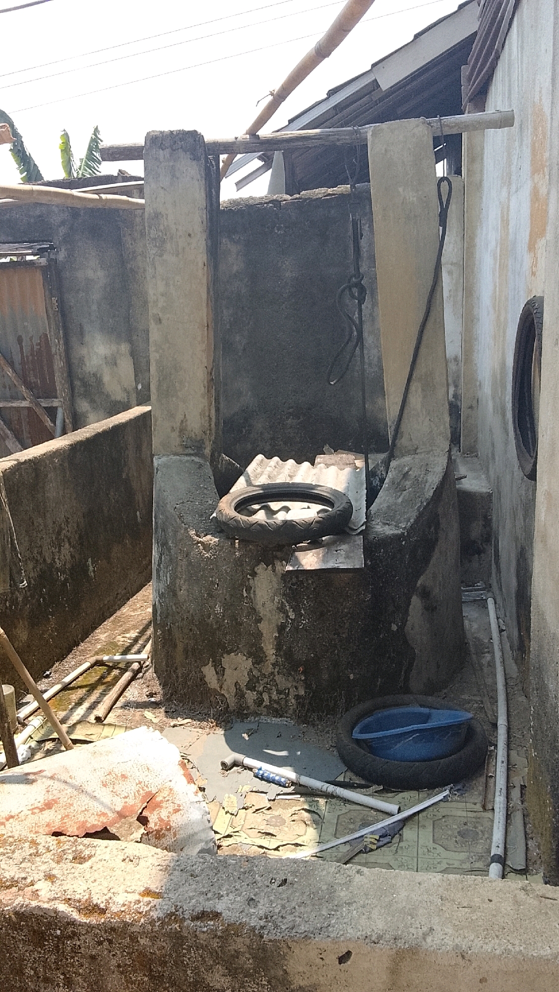 The image shows an outdoor scene in Bojong Barat, Indonesia, with a focus on a well structure and its surroundings. The well is made of concrete and appears aged, with some cracks and discoloration. Inside the well's structure, a car tire is placed on a piece of corrugated metal, and a blue plastic basin is also visible. In the foreground, there's a concrete ledge or wall, and to the left, a section of a larger concrete structure with visible rebar. The ground is covered with debris and what appears to be old tiles. Various pipes and wooden structures are scattered around the area, suggesting a functional or former utility area. The background shows a tiled roof and some vegetation, indicative of a residential or semi-rural setting. The lighting suggests it is daytime, with clear skies. There are no people visible in the image.