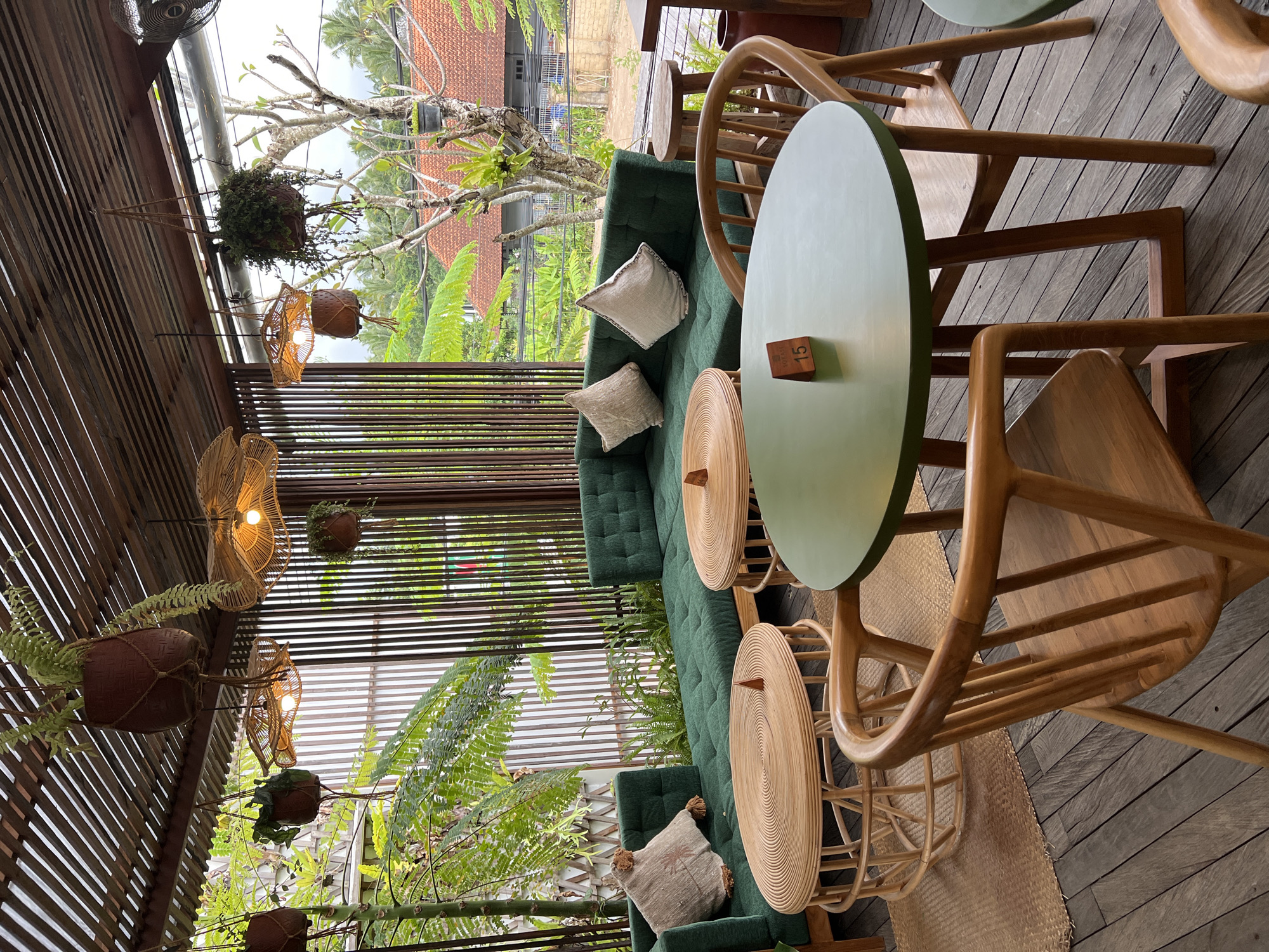 The photograph captures an outdoor seating area in Singakerta, Indonesia. The scene is set on a wooden deck. A long, green cushioned bench runs along the length of the area, adorned with several light-colored pillows. Two small, round wicker tables sit adjacent to the bench, each paired with two wooden chairs. The tables have a distinctive green top. A wooden block with the number "15" is placed on one of the tables. Overhead, a slatted wooden roof structure supports hanging plants in woven pots and decorative rattan light fixtures. Lush greenery, including ferns and other plants, surrounds the seating area, adding to the tropical ambiance. Through the slats in the roof, one can see buildings in the distance.