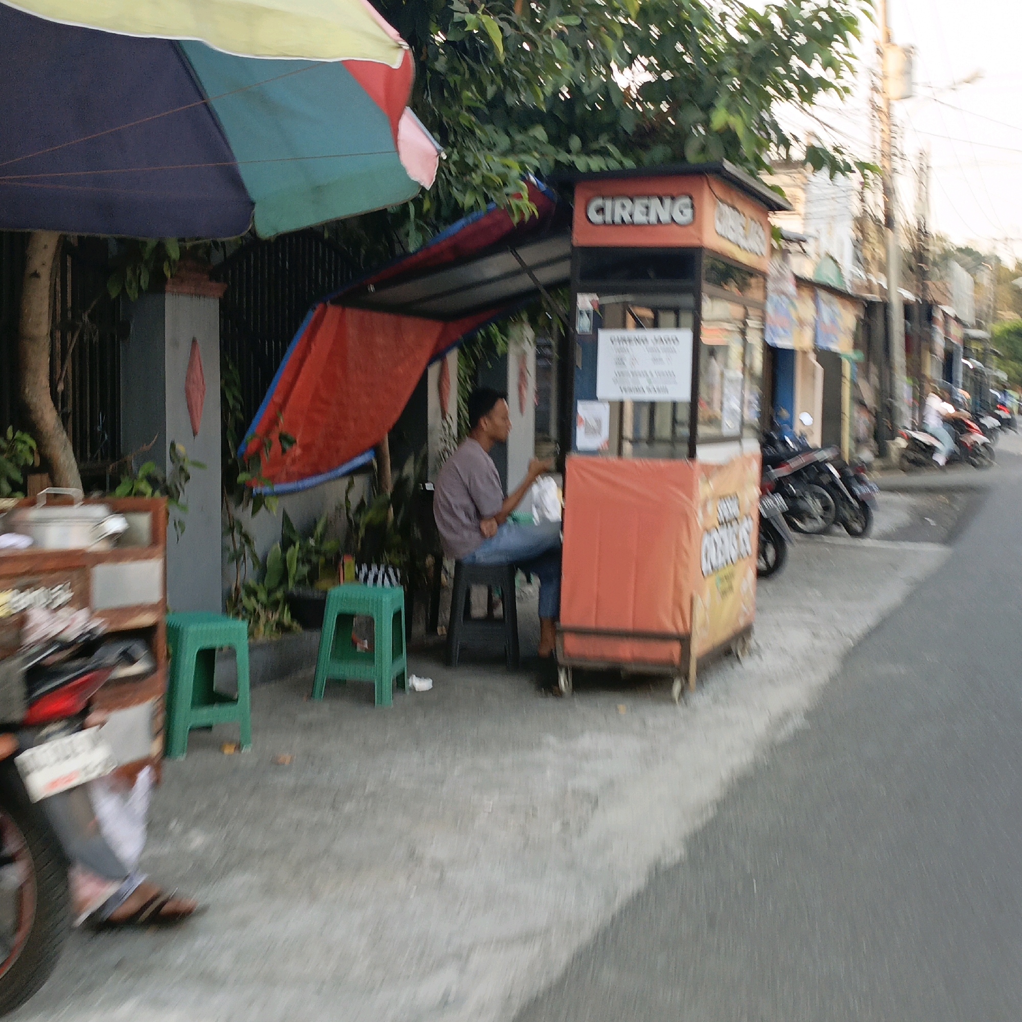This is a medium-shot, street-level, slightly blurred, candid photograph taken outdoors in what appears to be a residential or commercial area in Salatiga, Indonesia. The scene depicts a roadside food stall with a colorful umbrella overhead and a person sitting on a stool.
The food stall itself is constructed of wood and metal, with a bright orange tarp covering the front. Above the tarp, a sign with the word "CIRENG" in white capital letters on an orange background is visible. To the right of the "CIRENG" sign, another sign is partially visible. Behind the stall, a red tarp is draped, and a wooden cart with cooking utensils is to the left of the stall. Two green plastic stools are placed in front of the stall.
A man, wearing a grey t-shirt and blue jeans, is seated on a black stool, facing away from the camera and towards the stall. He appears to be holding something in his right hand. The background shows trees and foliage, a fence, and other buildings and street elements typical of an urban environment in Indonesia. A motorcycle is parked to the left of the stall, with its rear visible. Further down the street, other motorcycles and people are visible, suggesting a busy street scene. The ground is paved with concrete. The overall lighting suggests it is either early morning or late afternoon, with soft sunlight filtering through the trees.