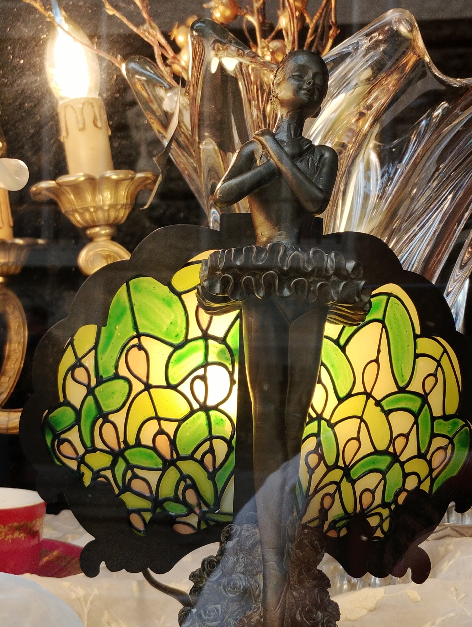 The image presents a detailed, dark metallic sculpture of a ballerina or female figure centrally positioned within an ornate indoor display. The figure has a serene expression, with hands clasped near her chest, and wears a ruffled skirt resembling a tutu.
Behind the sculpture is a decorative lamp featuring a scalloped black frame and a stained-glass-like shade. The shade displays an intricate mosaic pattern of interconnected green and yellow translucent pieces, illuminated from within to cast a warm glow.
To the upper left, an electric candle bulb, set in a golden candelabra-style holder, emits a bright, warm light. Further back and to the right of the ballerina, a clear, possibly glass or crystal, decorative object with undulating, reflective surfaces adds to the elaborate aesthetic. Hints of dried branches or floral elements are visible behind these items.
In the bottom left corner, a portion of a red cup adorned with gold detailing is visible. The foreground and bottom right show white fabric, possibly a tablecloth or draped material. The overall scene suggests an indoor setting, such as a shop window display or home decor, characterized by artificial lighting, implying an evening or low-light environment. The arrangement evokes a sense of classical elegance and decorative artistry. No text is visible.