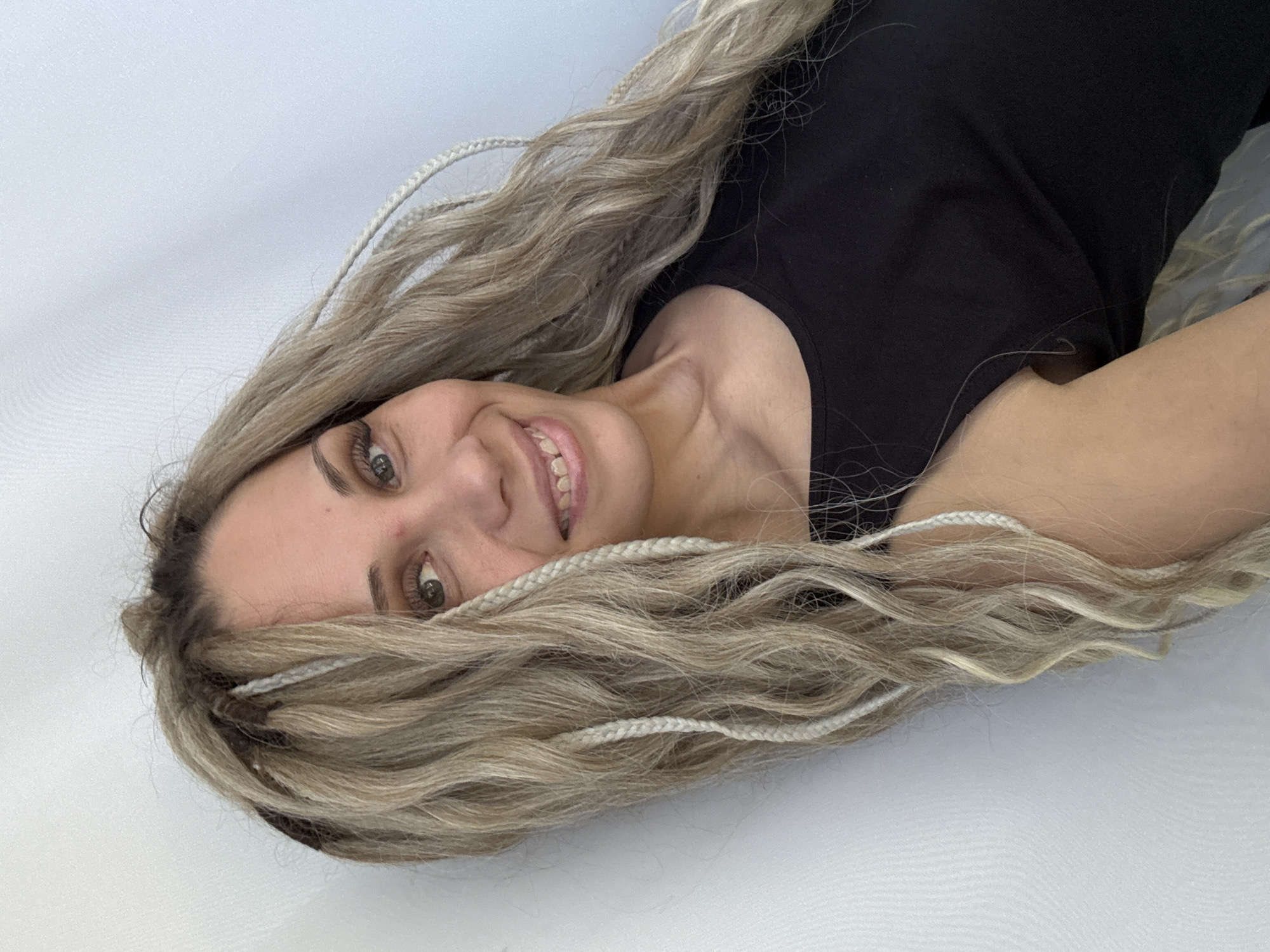 A fair-skinned woman with long, wavy blonde hair interspersed with white braids is smiling and looking directly at the camera. She's wearing a black T-shirt. The background appears to be a plain white surface. There is no indication of the image being taken in Rusanovo, Russia, or any other specific location.