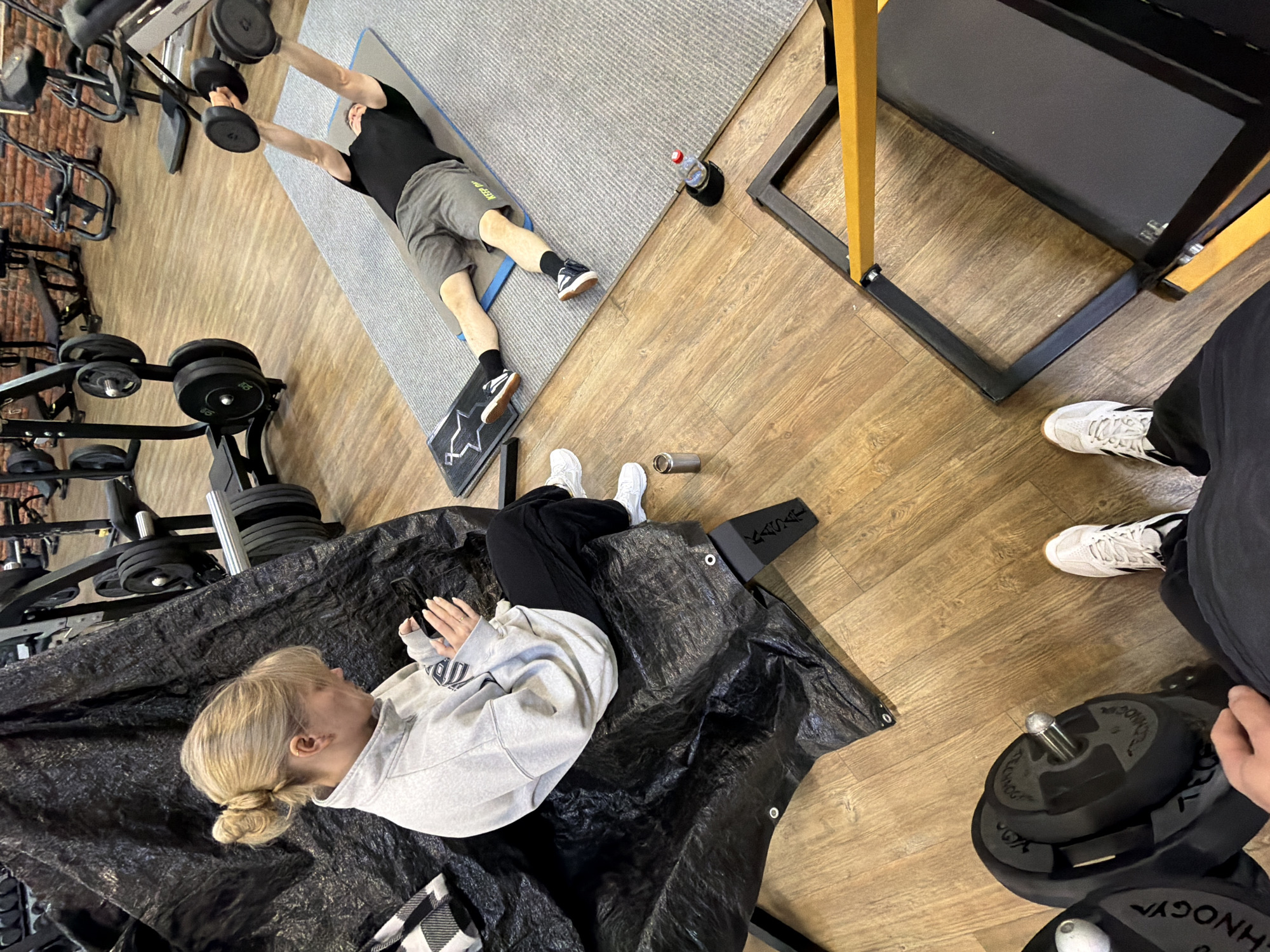This overhead image captures an indoor fitness session at a gym in Chaikovsky, Russia. The setting features a light-toned wood laminate floor and various pieces of workout equipment.
In the mid-ground, a man with short dark hair, wearing a black t-shirt, grey shorts with "KEEP DO" partially visible, black socks, and white sneakers, lies on a grey exercise mat. He is actively performing an exercise, holding black dumbbells overhead with outstretched arms. His feet rest on a small black mat adorned with a white star-like design. Near his head, a red-capped water bottle sits in a black holder.
In the foreground, a woman with blonde hair styled in a bun sits on a large black tarp or protective sheet. She is dressed in a light grey hooded sweatshirt, which appears to have a logo (possibly "UM") on the front, black pants, and white sneakers. She is looking towards the exercising man, holding a smartphone, suggesting she is observing, resting, or taking a break. A silver metallic bottle lies on the floor near her feet. Partially visible beneath her is a black and white checkered fabric item.
To the right, the legs and feet of a third person, wearing black pants and white sneakers with black stripes, are visible. Nearby, a weight rack holds several black weight plates, some marked with "VAGYGYM." A large black and yellow metal frame, likely part of a squat rack or similar equipment, is also visible on the right side of the frame. The scene depicts a focused and active gym environment.