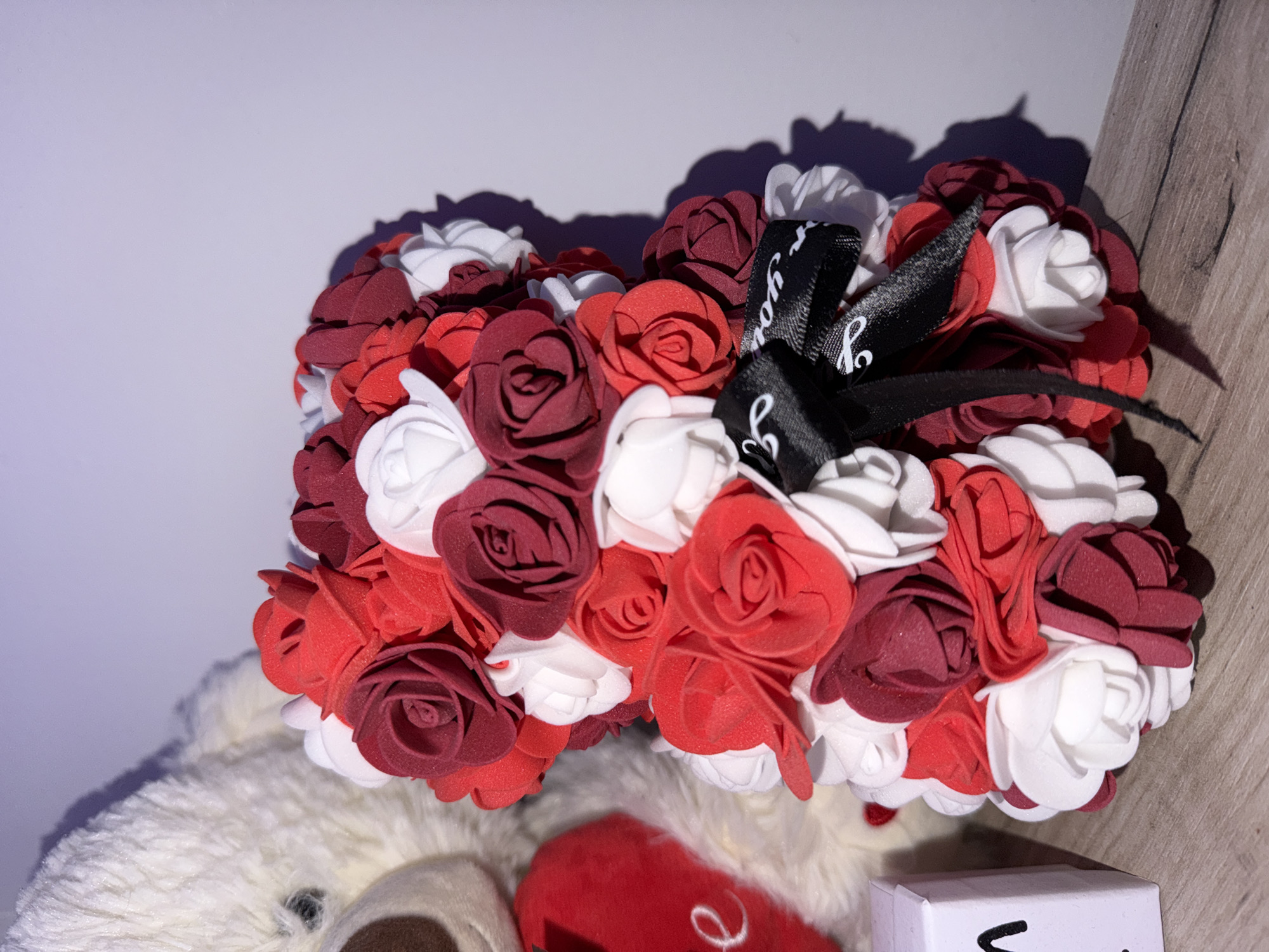 An indoor scene displays decorative items on a wooden surface against a plain, light-colored wall. The central object is a teddy bear figure composed entirely of artificial roses in red, maroon, and white hues. A black ribbon tied into a bow, featuring white cursive text, is affixed to the bear's chest area. To the left, a portion of a larger, light-colored plush teddy bear with a brown snout is visible. Adjacent to this plush bear, a red heart-shaped object, partially showing a white cursive "e," is present. Further left, a small, white, rectangular object with black markings is partially discernible. The items are illuminated by an artificial light source, casting distinct shadows onto the wall and the wooden surface. The specific location within the city of Varna, Bulgaria, cannot be determined from the visual evidence.