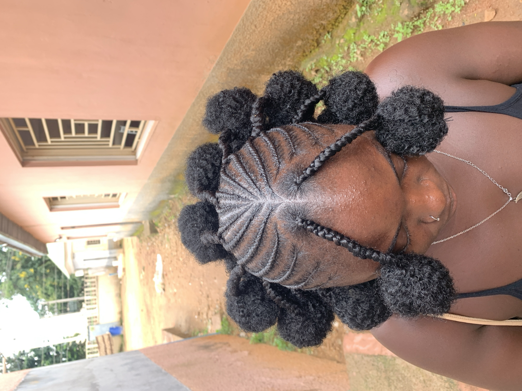 The image shows a close-up of a young Black woman's head from a high angle, as she looks down. Her hair is styled in intricate braids that radiate from a central part, with each section of hair twisted into a round, voluminous puff. Some of the braided sections hang down as individual braids with similarly styled puffs at the ends. She is wearing a black tank top and a silver necklace with a pendant.
The background appears to be an outdoor setting. To the right, a peach-colored building with barred windows is visible. To the left, a gray and light brown wall runs parallel to a dirt path. The ground is covered in dirt and some sparse green vegetation. The overall impression is of a residential area, possibly in a town or village in Nigeria. The specific location of New Stadium, Nigeria, is inferred from the provided information, but not directly observable from the image itself.